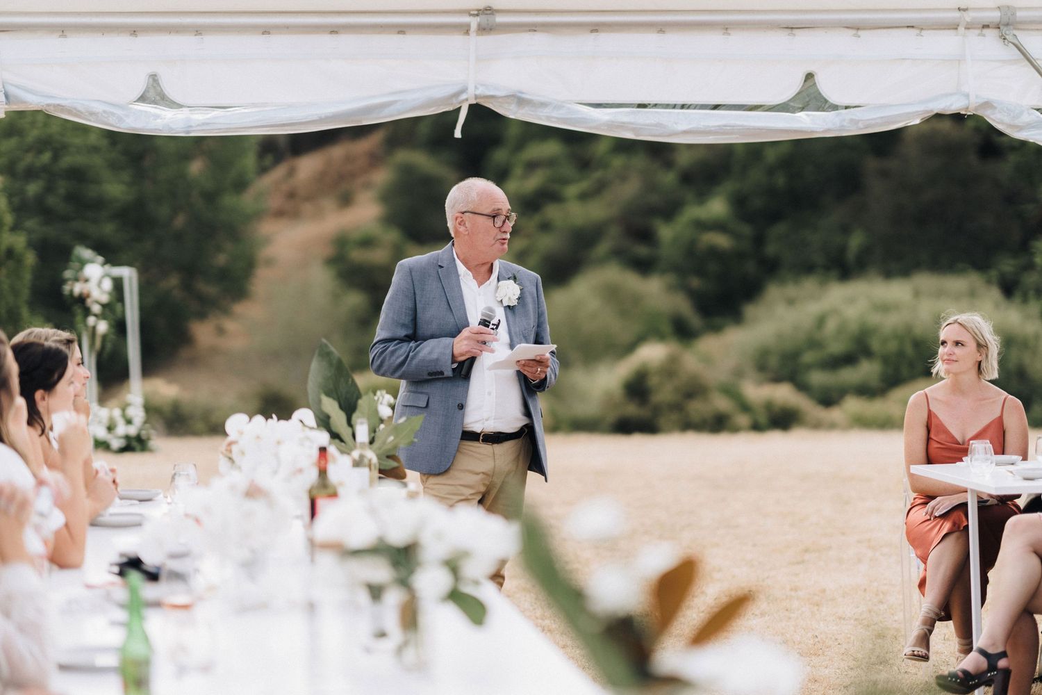 Wedding reception speech being given under a white tent canopy in an outdoor setting.