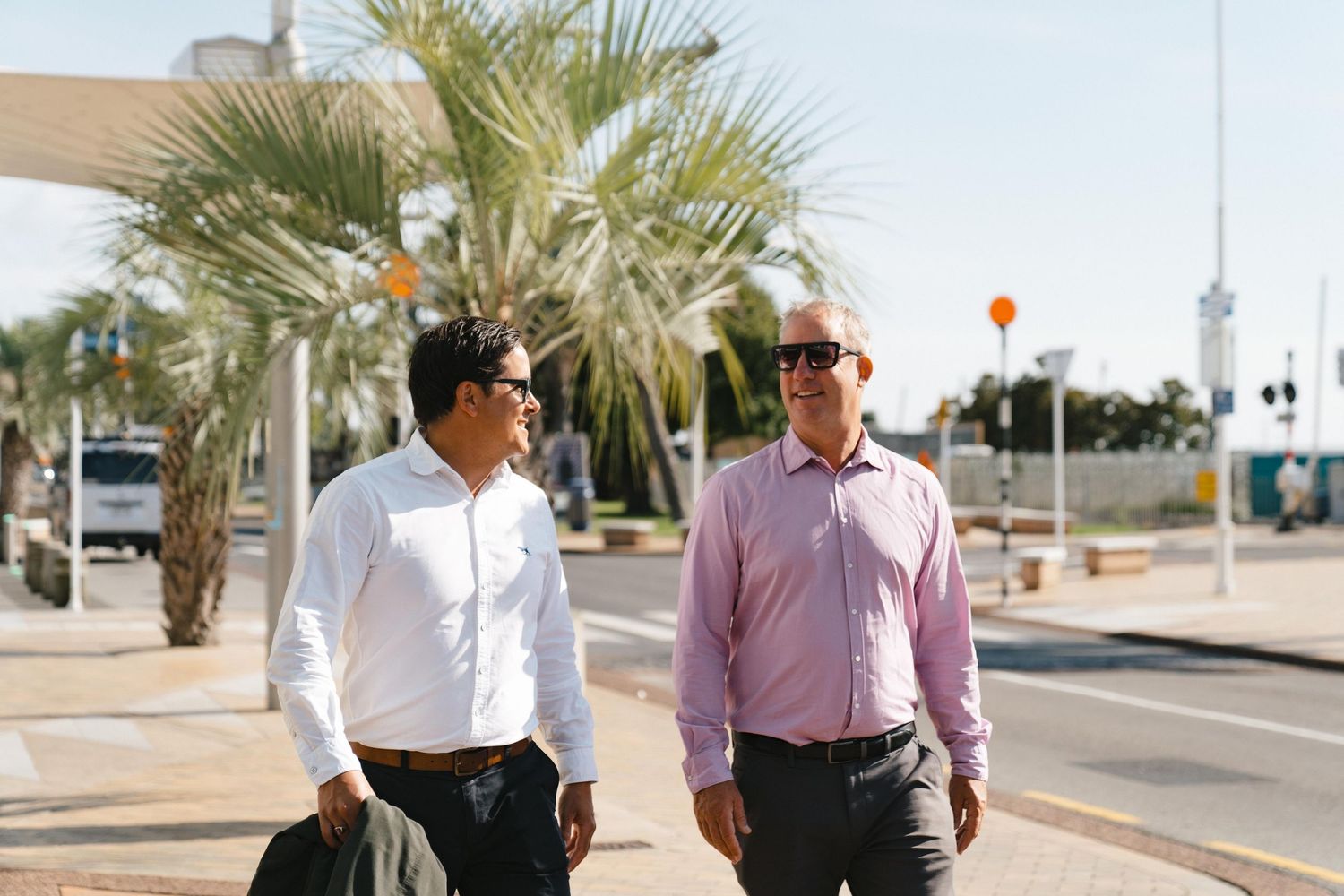 Two businessmen walking along a palm tree-lined street in business attire.