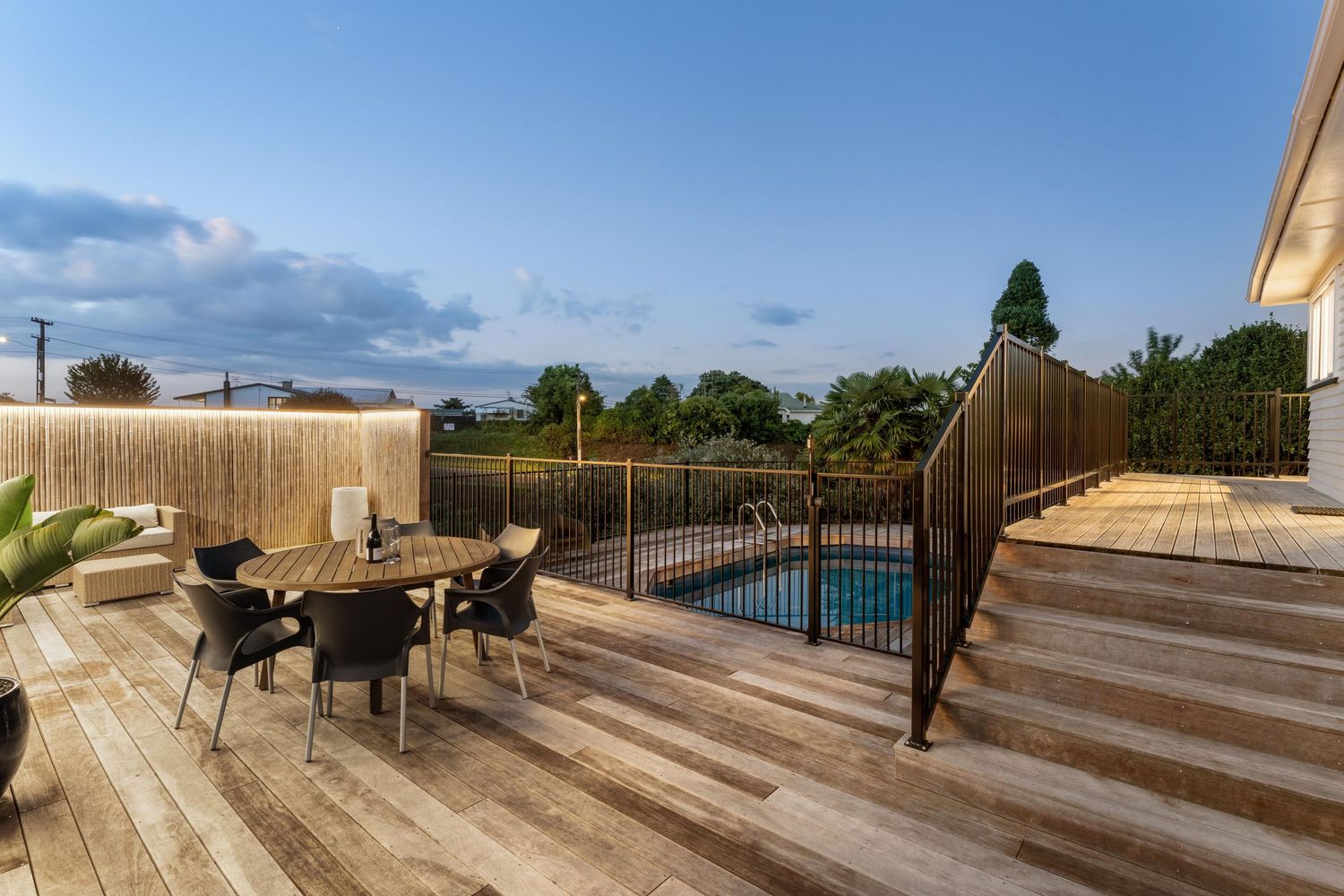 Large wooden deck with outdoor furniture overlooking a pool area under blue skies.
