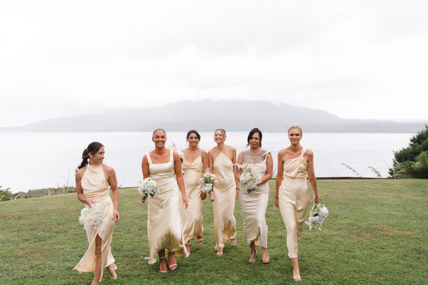A group of bridesmaids in champagne-colored dresses walk joyfully across a grassy hilltop with misty mountains behind them.