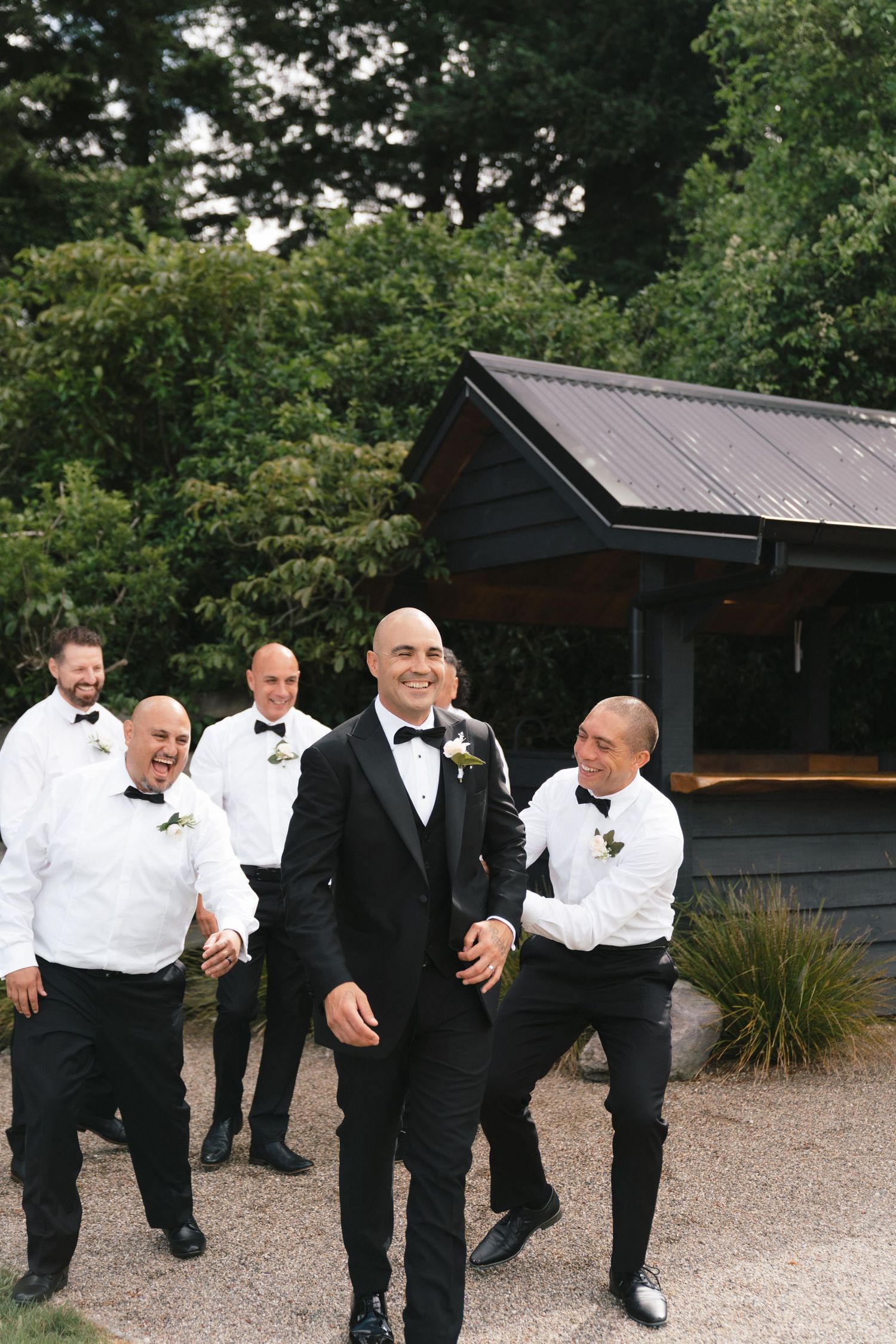 Groomsmen in black suits posing playfully on gravel path.
