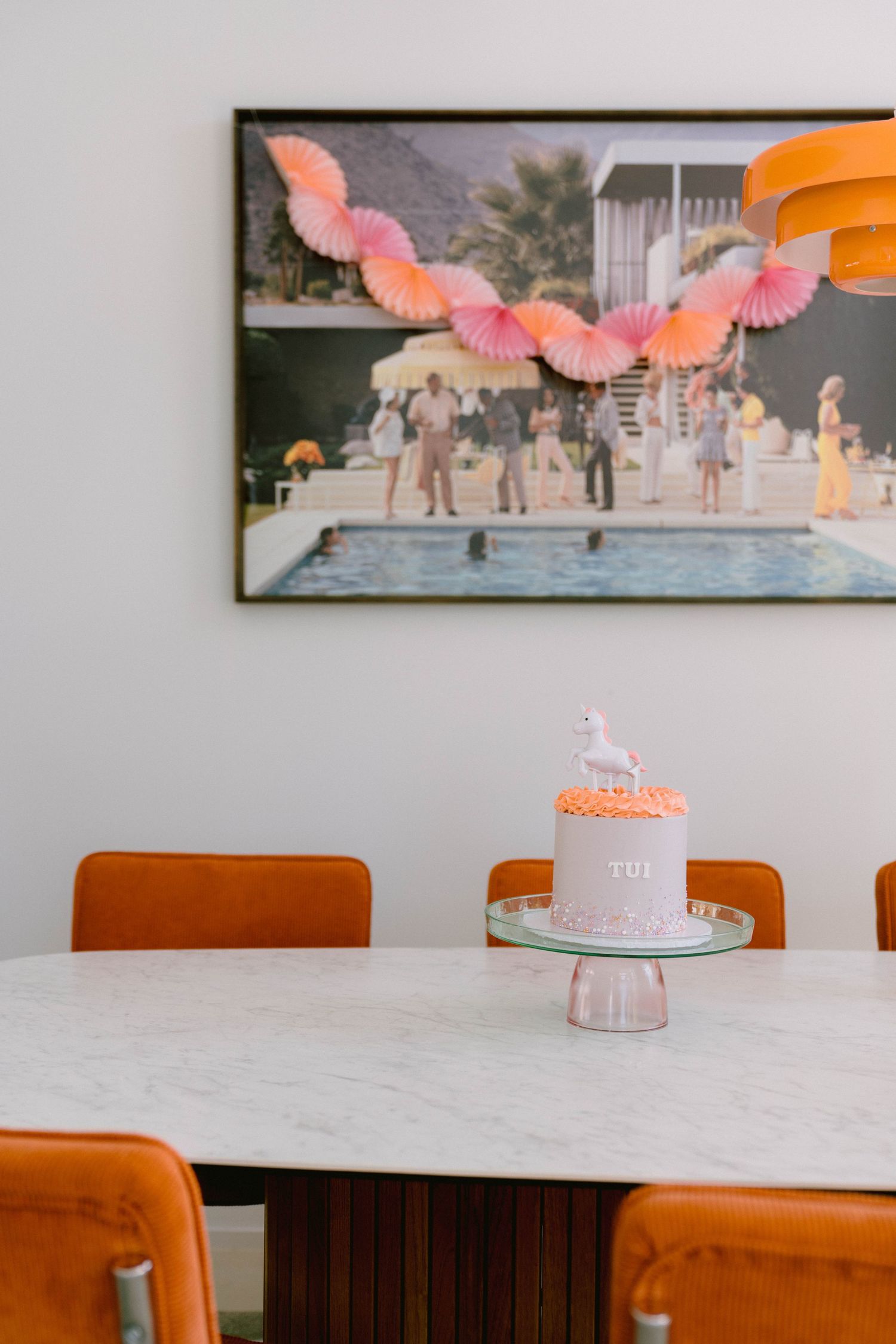 Retro-style dining room with orange chairs, marble table, vintage poolside artwork, and pink birthday cake on stand.