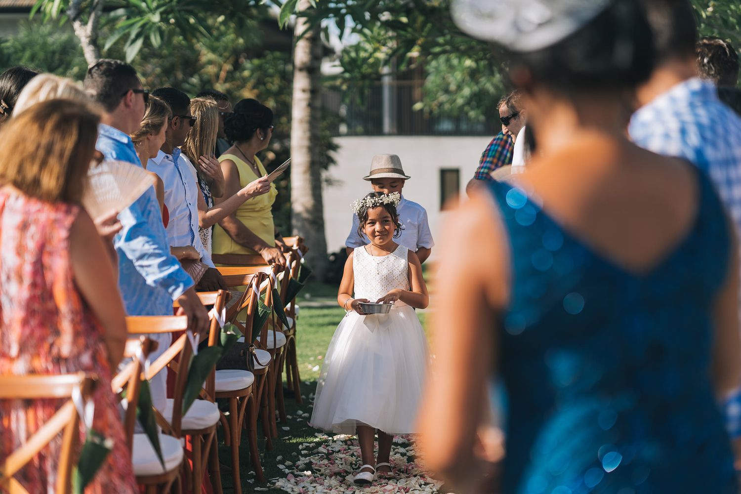 Young flower girl in white dress walks down outdoor wedding aisle surrounded by seated guests.