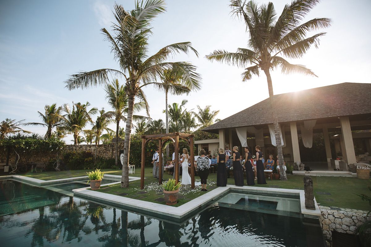 Elegant outdoor wedding ceremony setup with palm trees and infinity pool at tropical resort during sunset.