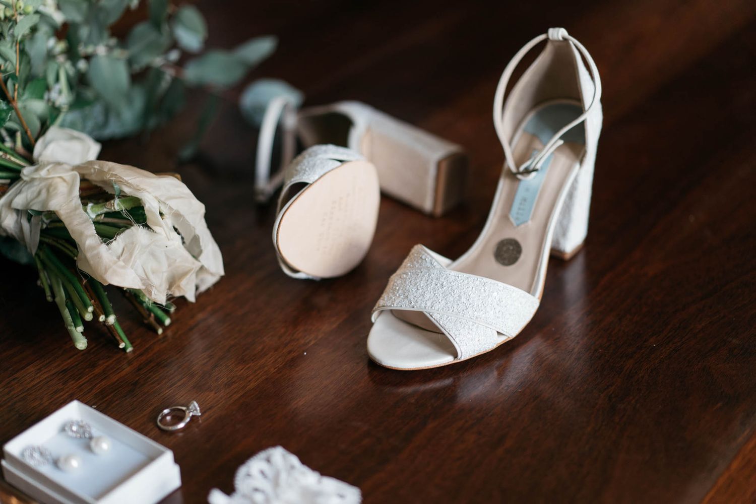 An elegant white bridal shoe with bow detail sits beside floral arrangements on a dark wooden surface.