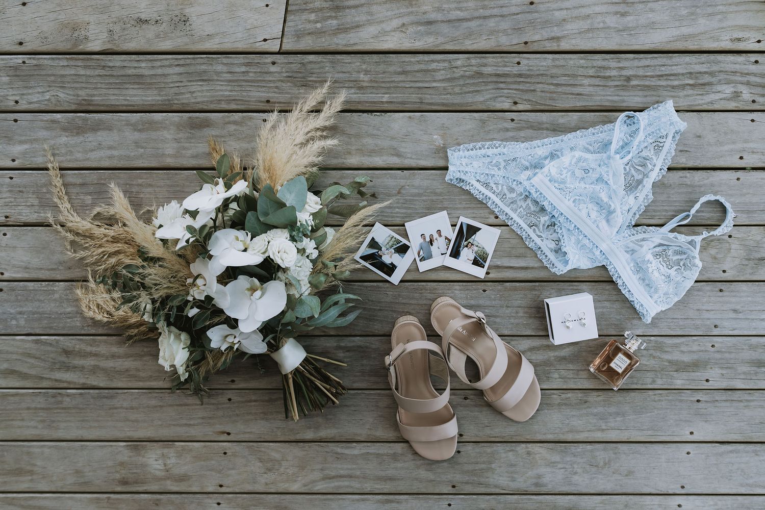 Wedding accessories including lace garter, bridal sandals, dried floral crown, and delicate white blooms arranged on wooden decking.