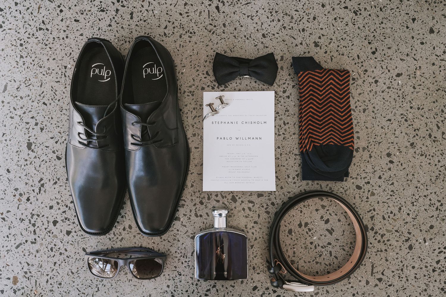 A flat lay arrangement of men's dress shoes, socks, sunglasses, bow tie, and accessories on a speckled gray surface.