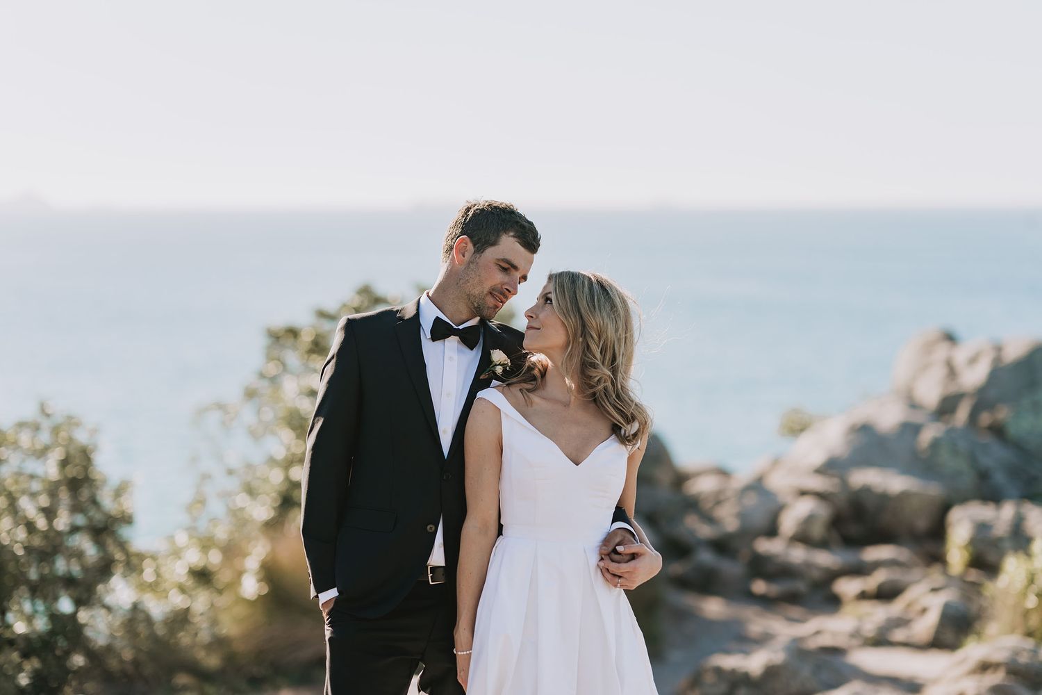 A couple in formal wedding attire embraces while standing on a scenic coastal cliffside during golden hour.