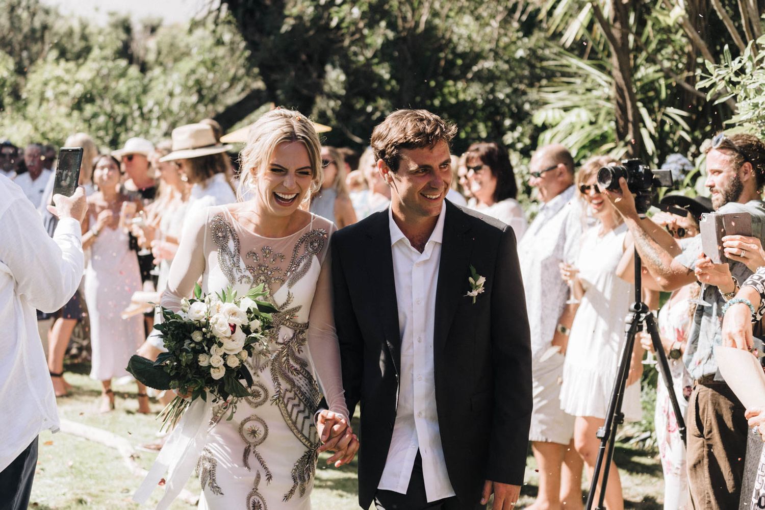 A newlywed couple walks down an outdoor aisle while guests celebrate with confetti in a sunny garden setting.
