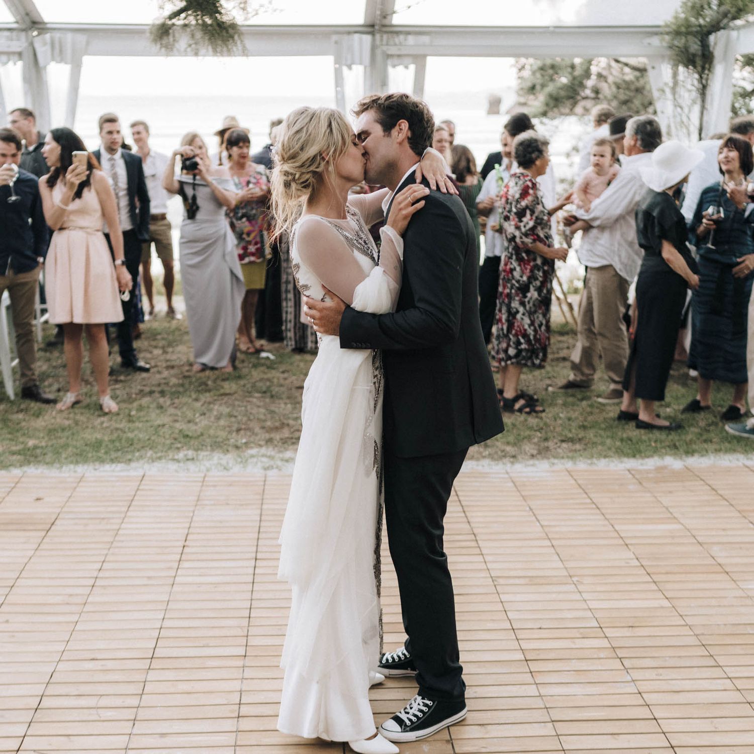 Newlywed couple shares their first dance on an outdoor tiled floor under a white tent while guests look on.