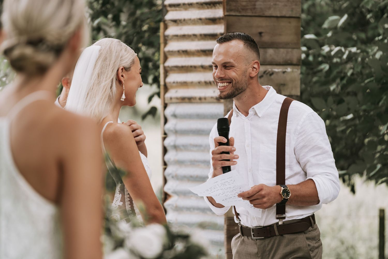 During an outdoor wedding ceremony, a groom in suspenders reads vows while holding a microphone under a rustic archway.