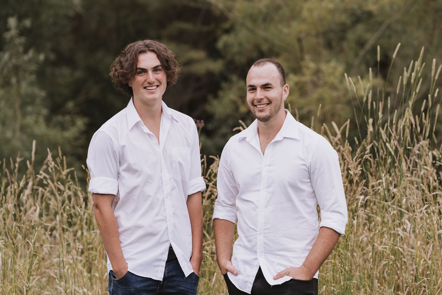 Two people wearing white button-down shirts stand together outdoors in tall grass with blurred natural background.