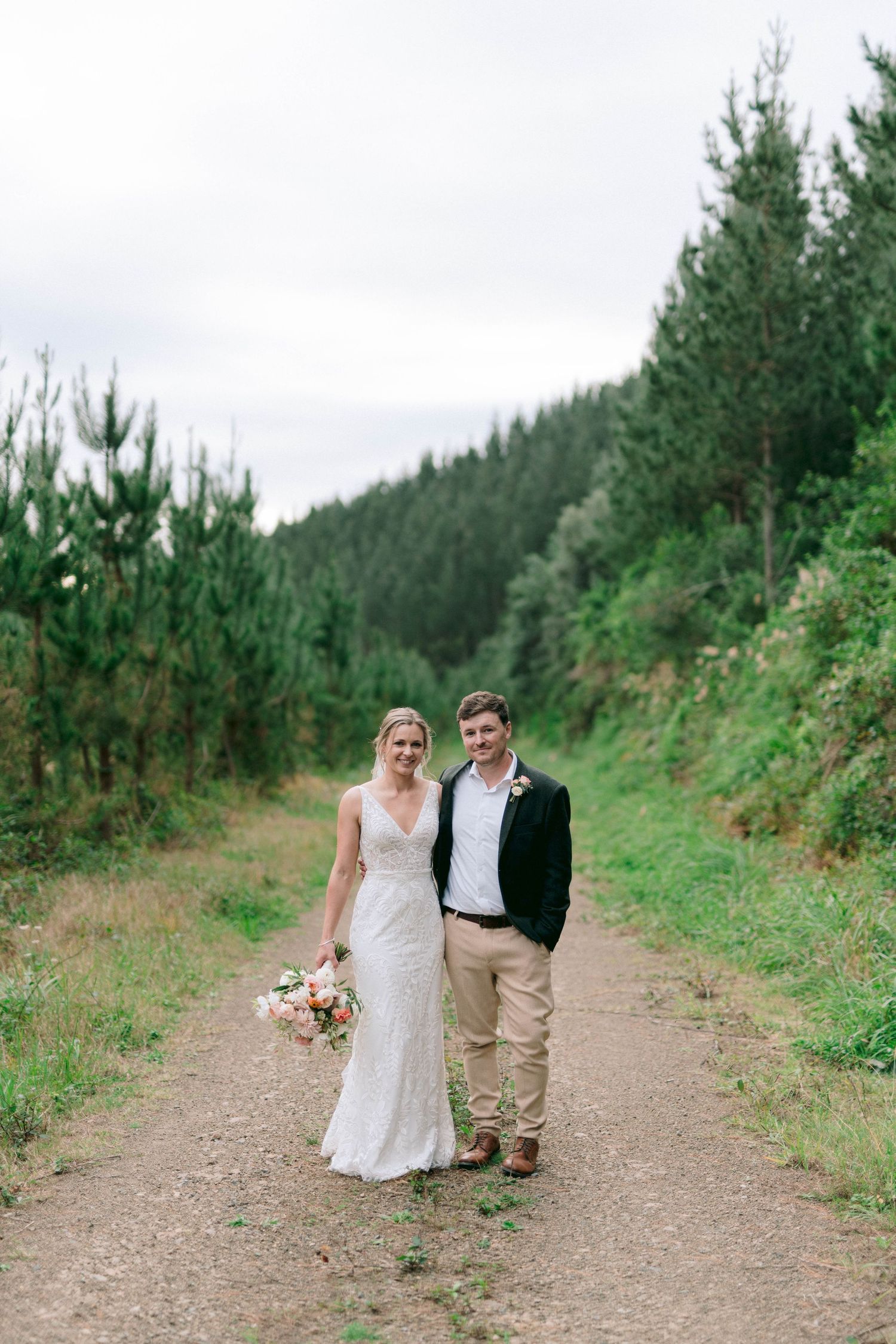 A couple poses together on a dirt path surrounded by evergreen trees in a scenic mountain setting.