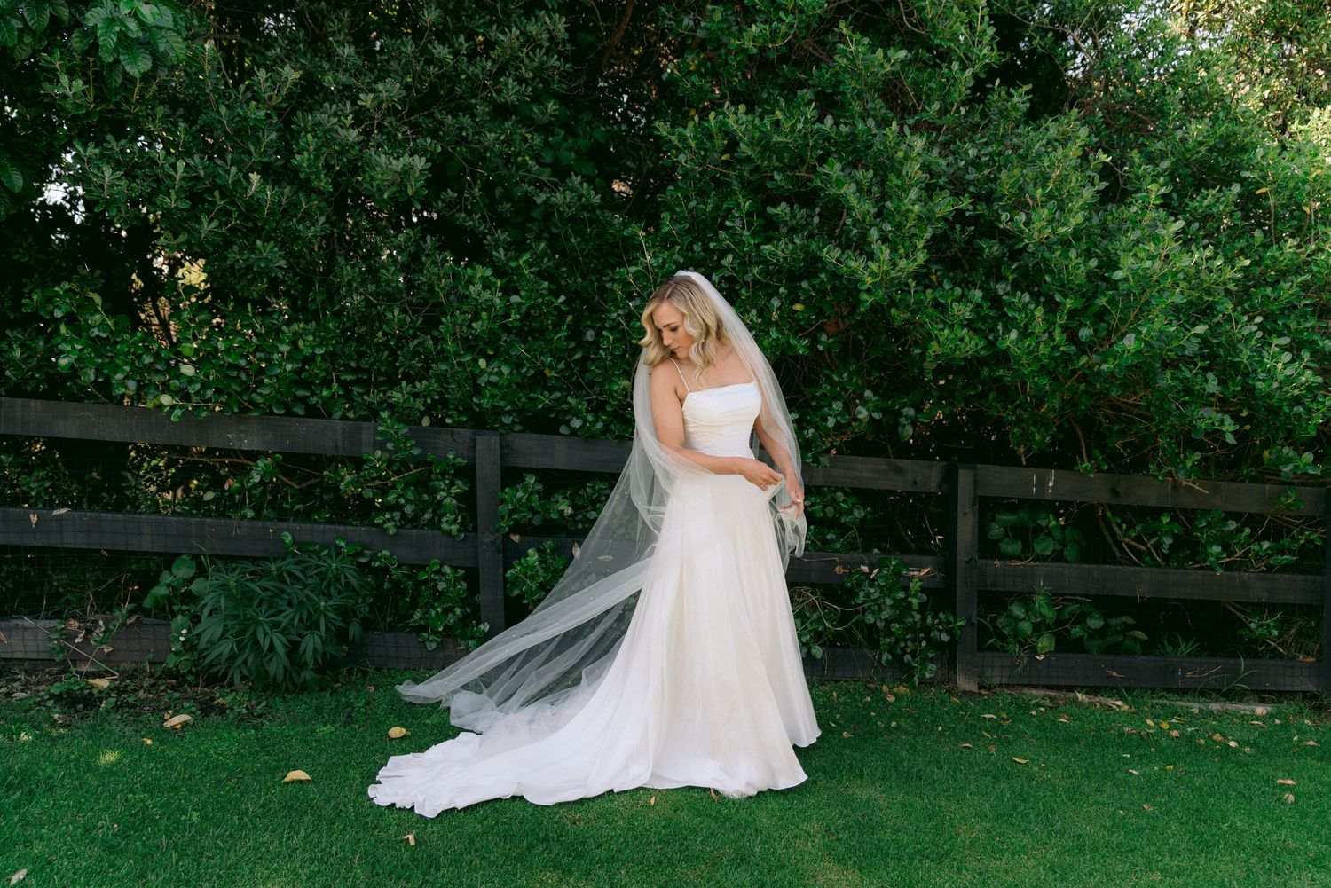 A bride in a flowing white wedding dress and veil stands on green grass near a wooden fence in a garden setting.