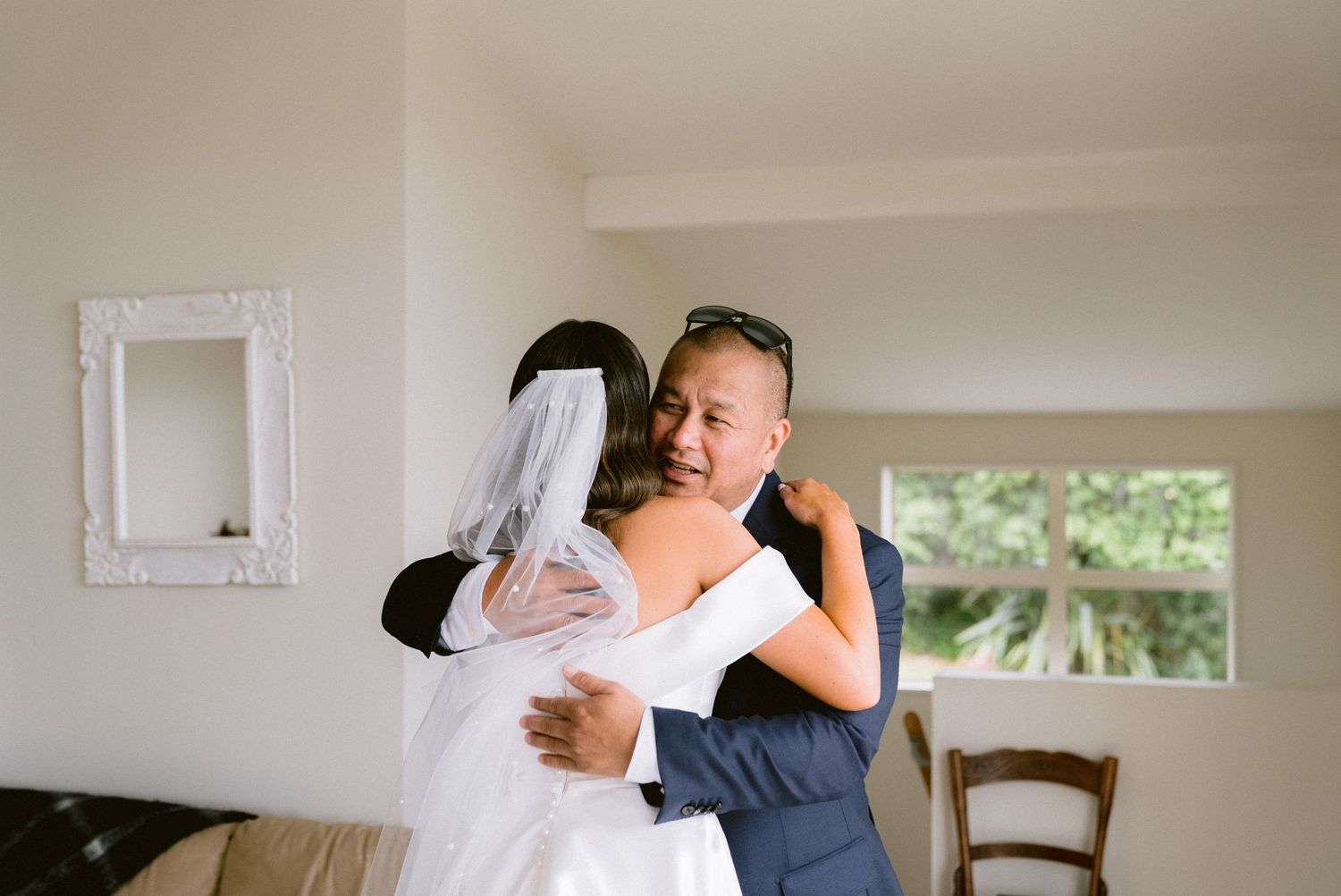 Two people share an emotional embrace in a bright room with white decor and a window view.