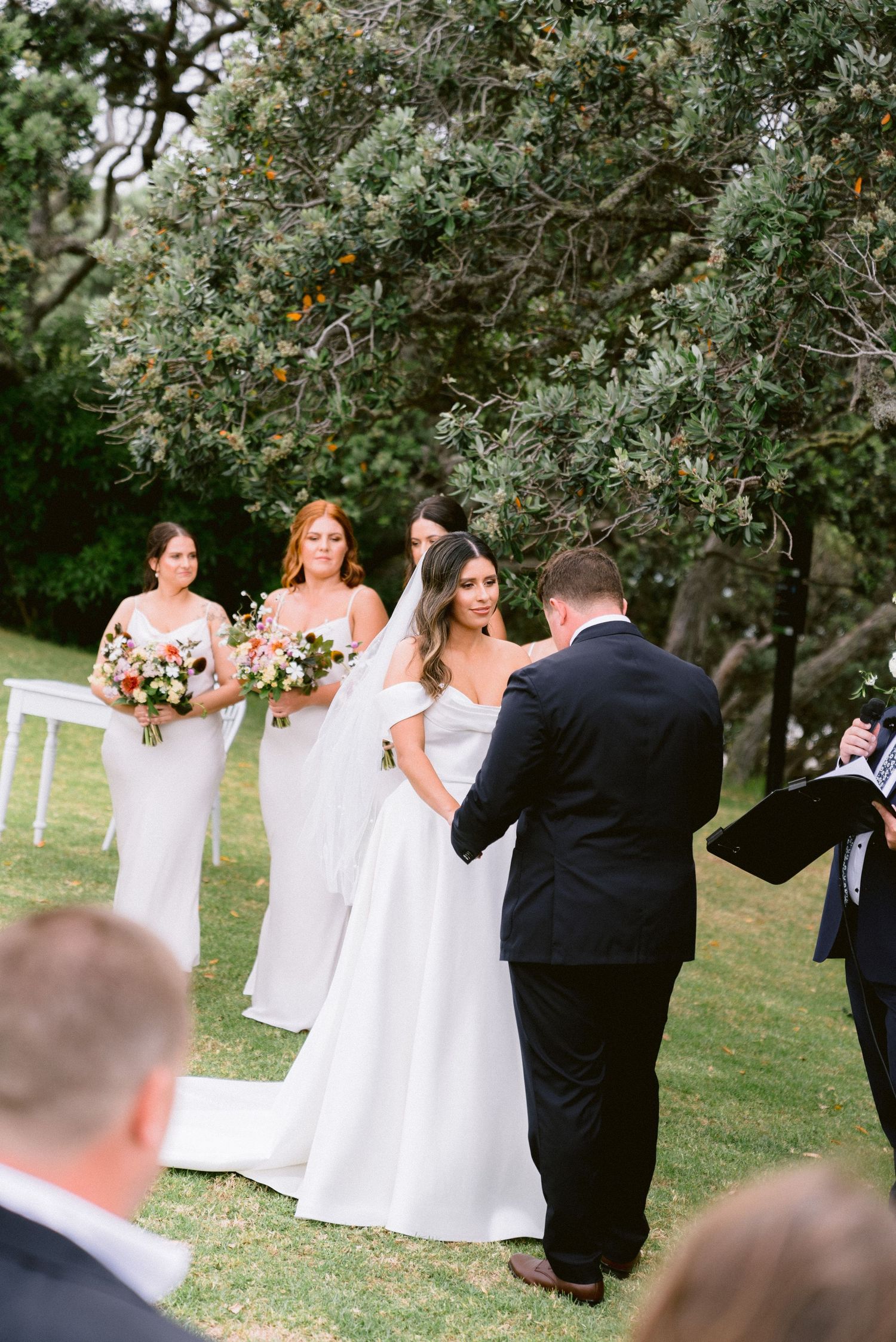 An outdoor wedding ceremony taking place under trees with bridesmaids in white dresses.