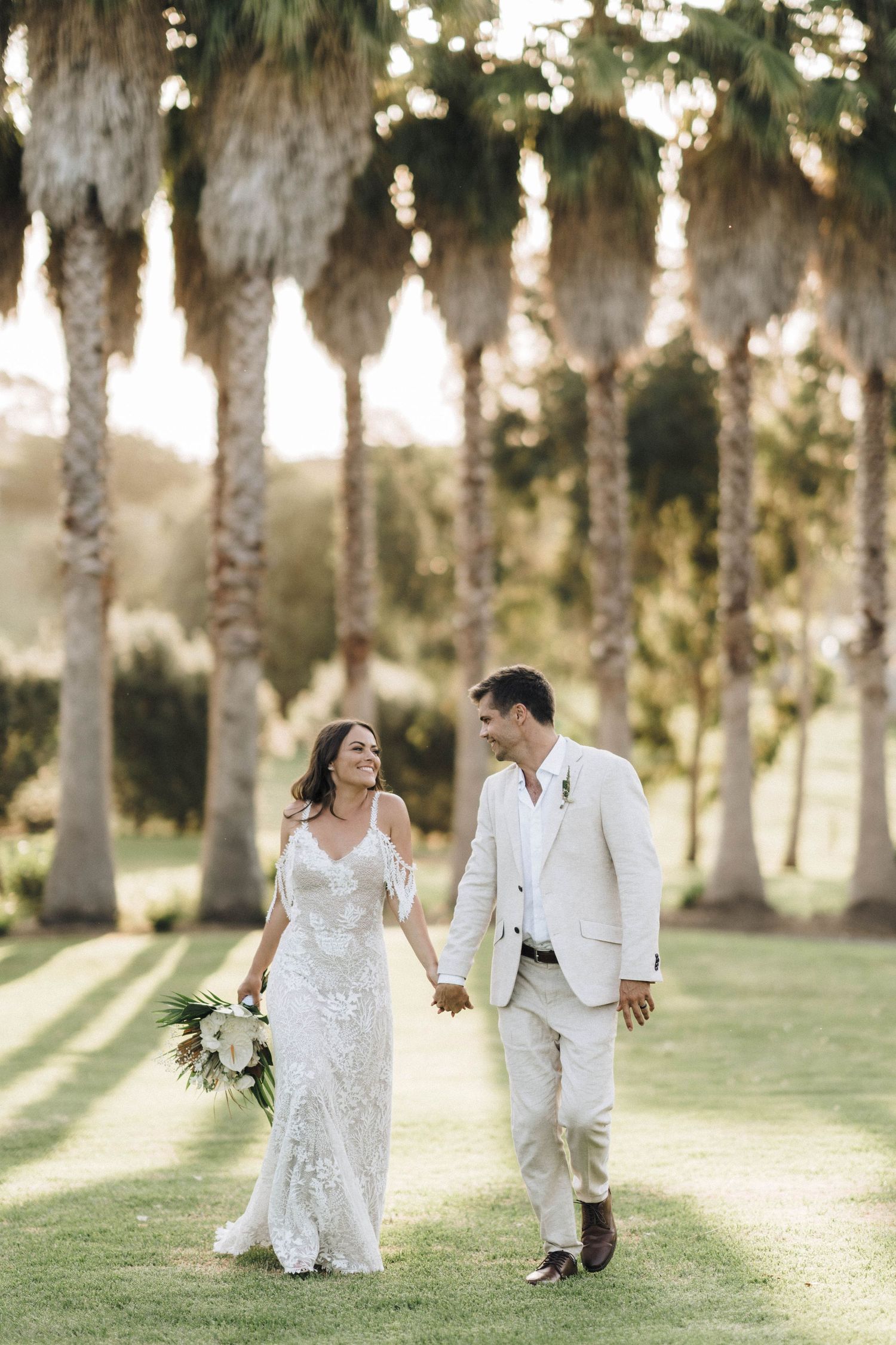 Couple walks together through a palm tree grove at sunset in elegant white wedding attire.