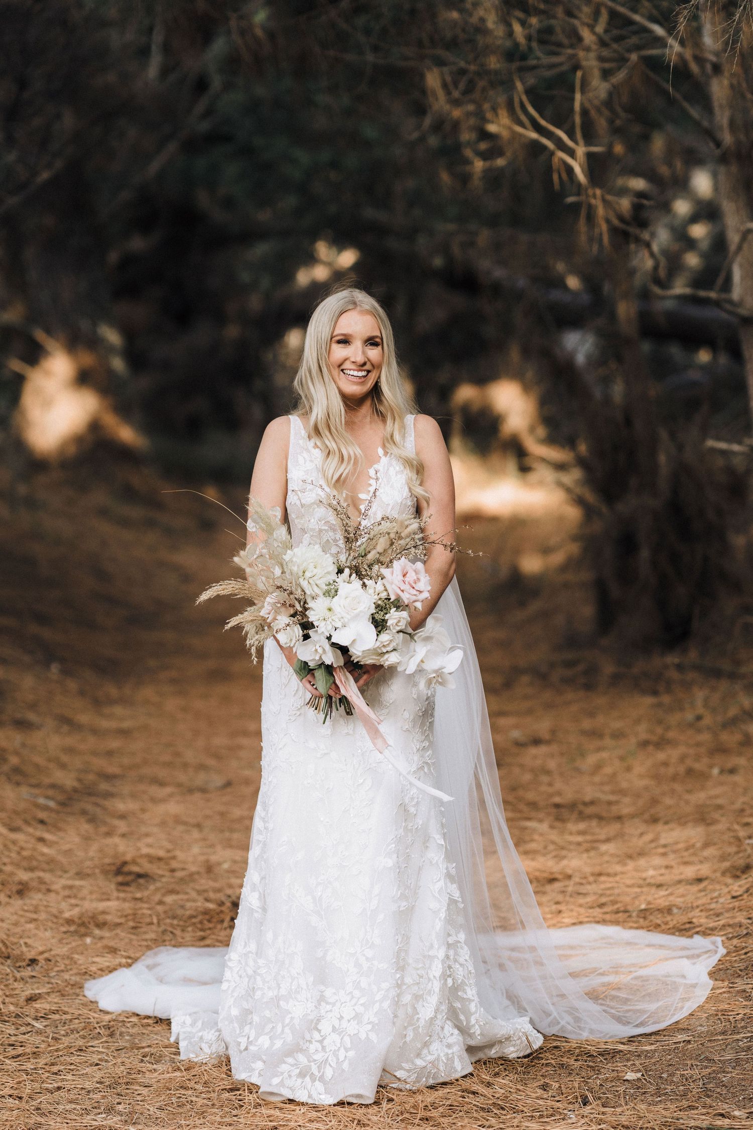 A bride in a lacy white wedding dress poses for photos outdoors on natural terrain during golden hour.