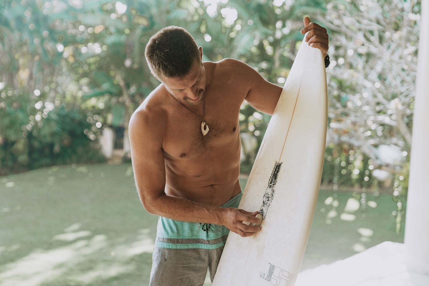 A shirtless person with mint green swim shorts inspects a white surfboard in a tropical outdoor setting.