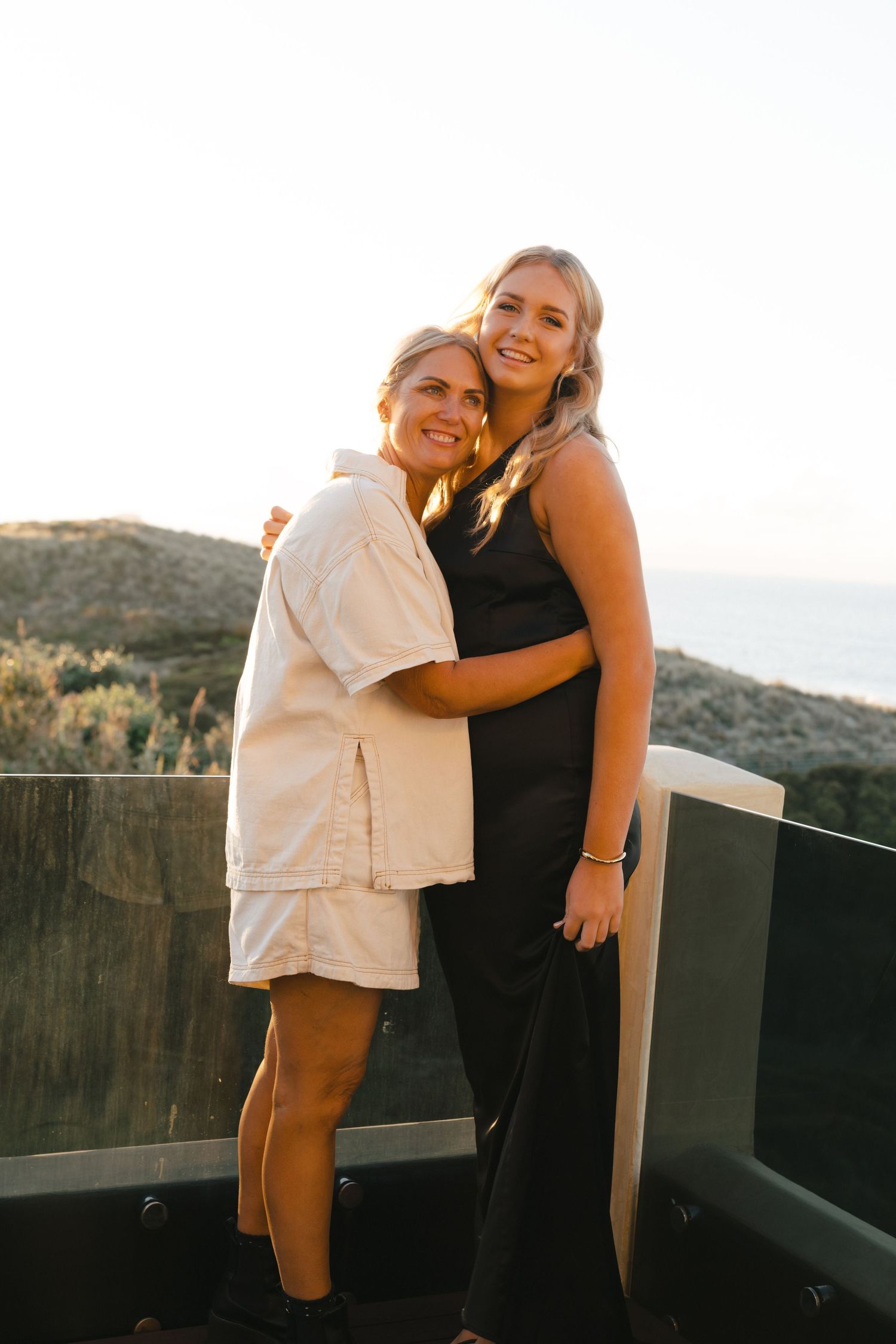 Two friends embracing and smiling together on an outdoor deck at sunset with mountains in the background.