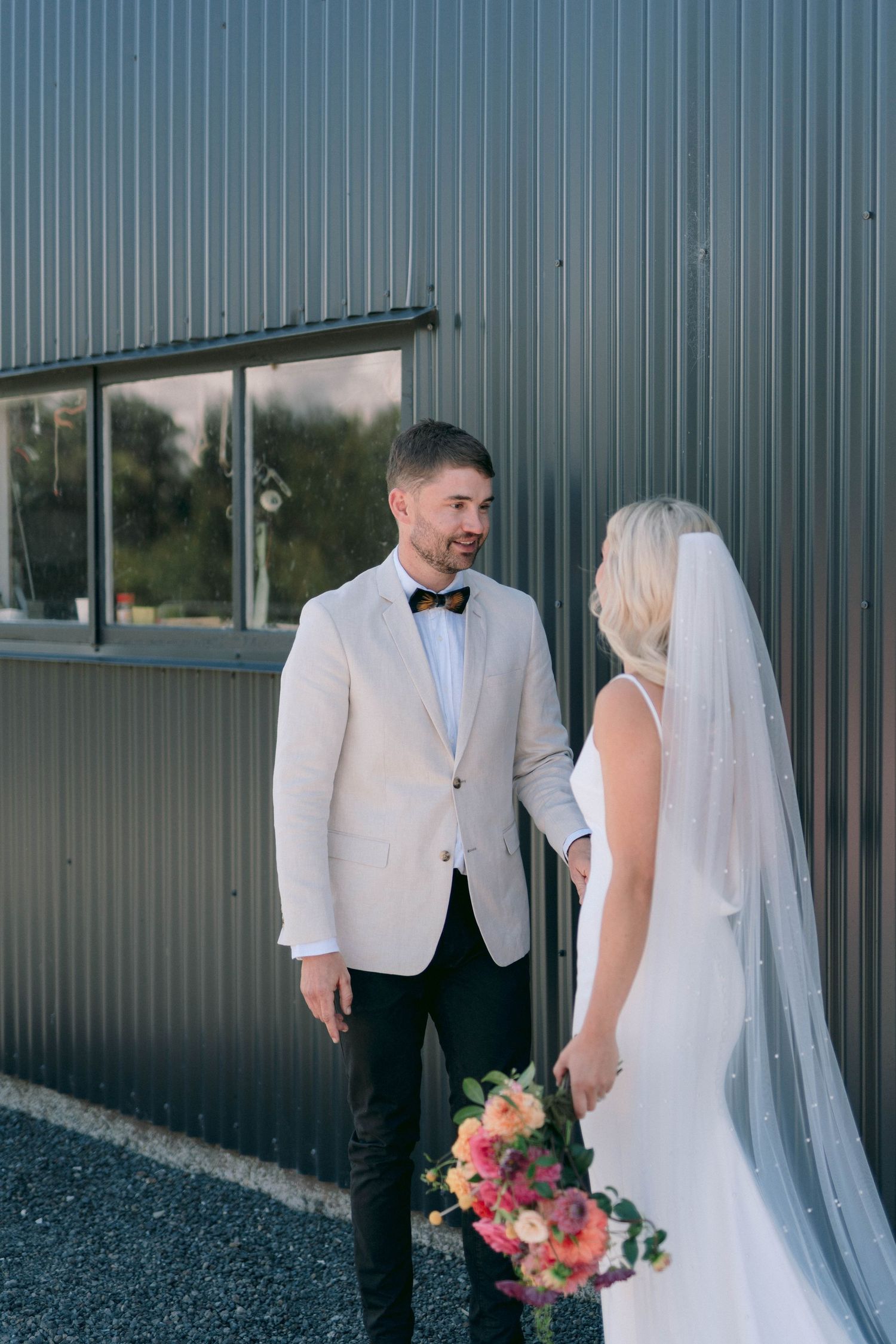 Newlyweds share a moment against an industrial wall as bride holds vibrant pink and coral bouquet.