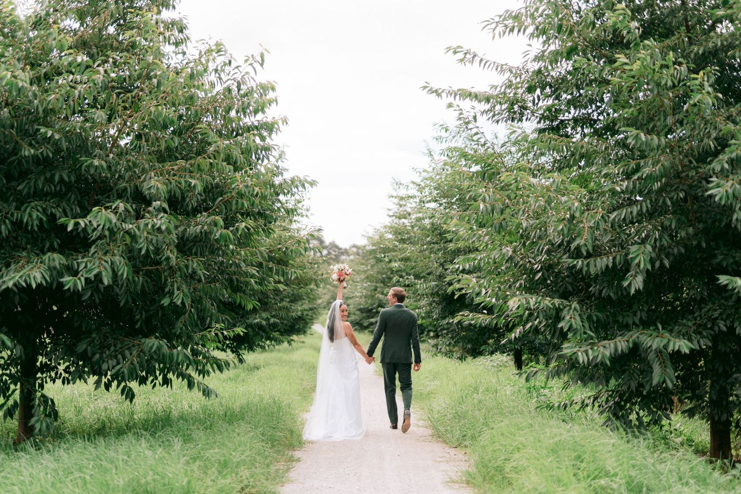 A wedding couple walks down a tree-lined path surrounded by lush green foliage on a bright summer day.