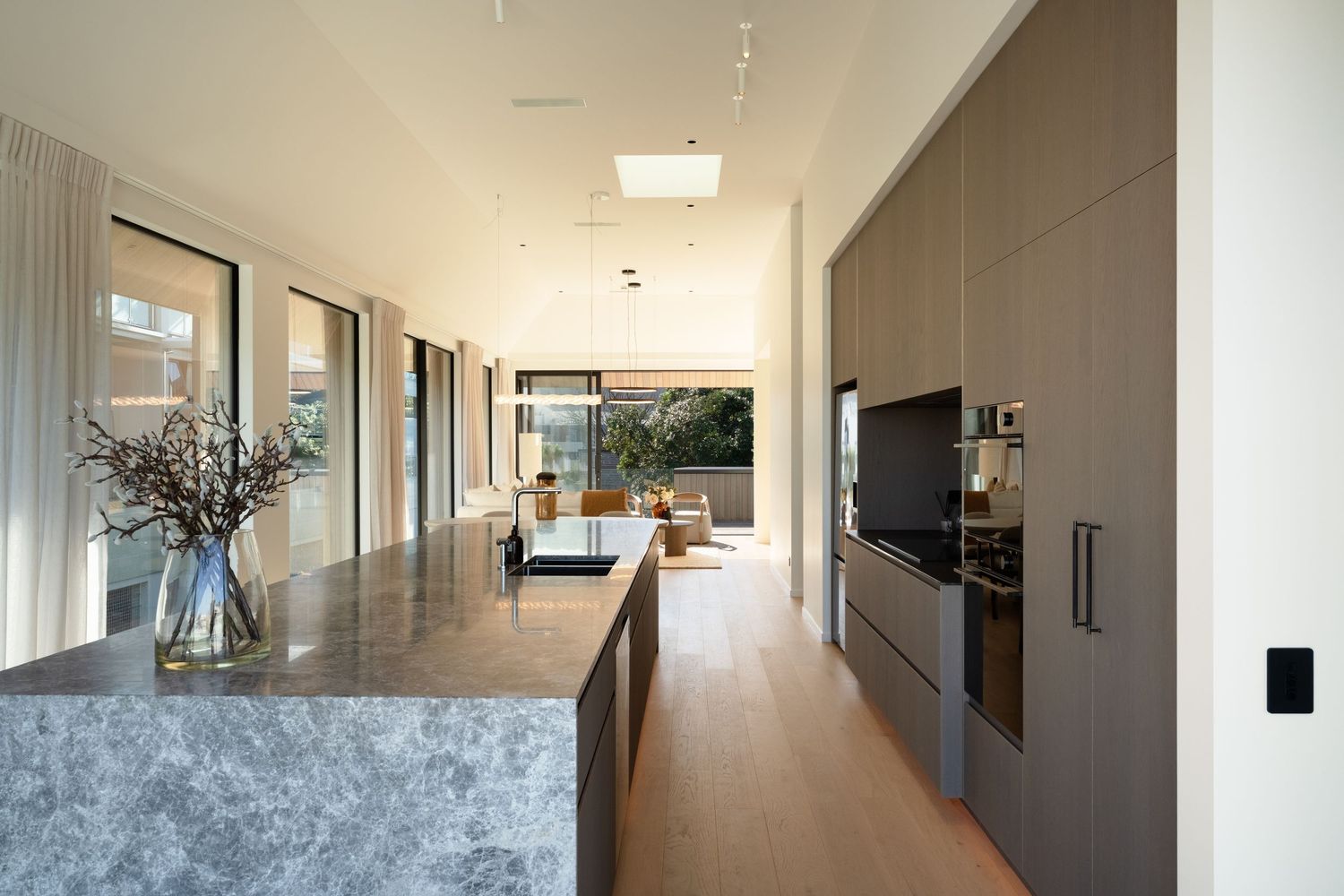 Modern open-plan kitchen with gray stone island, sleek cabinetry, hardwood floors, and floor-to-ceiling windows overlooking a courtyard.