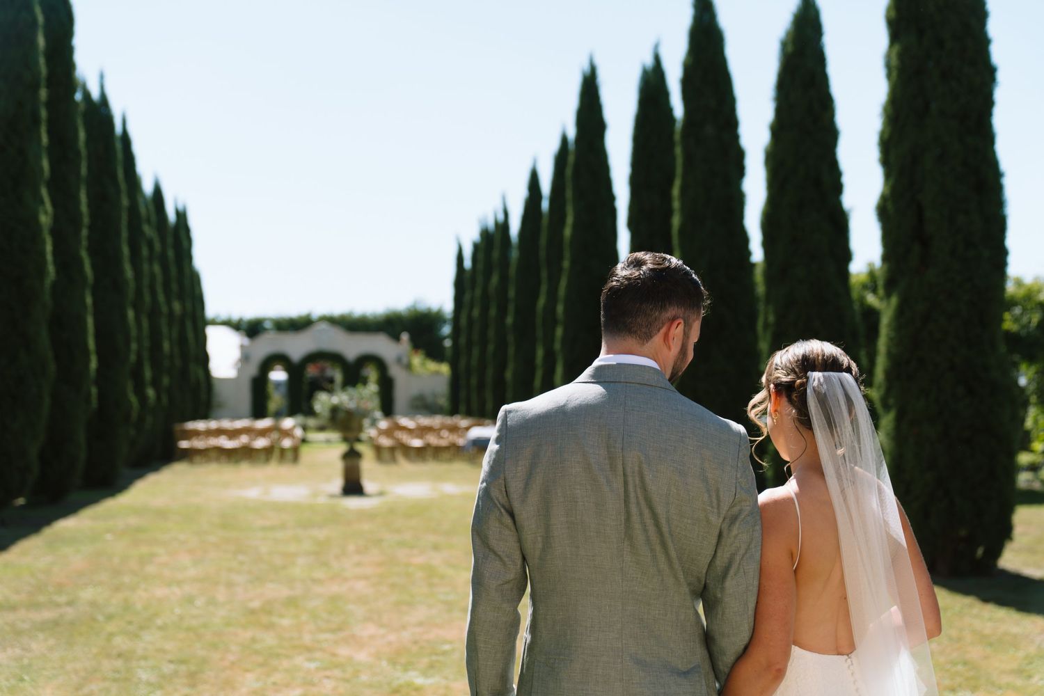 A couple in wedding attire walks down a cypress tree-lined path towards a villa in the Italian countryside.