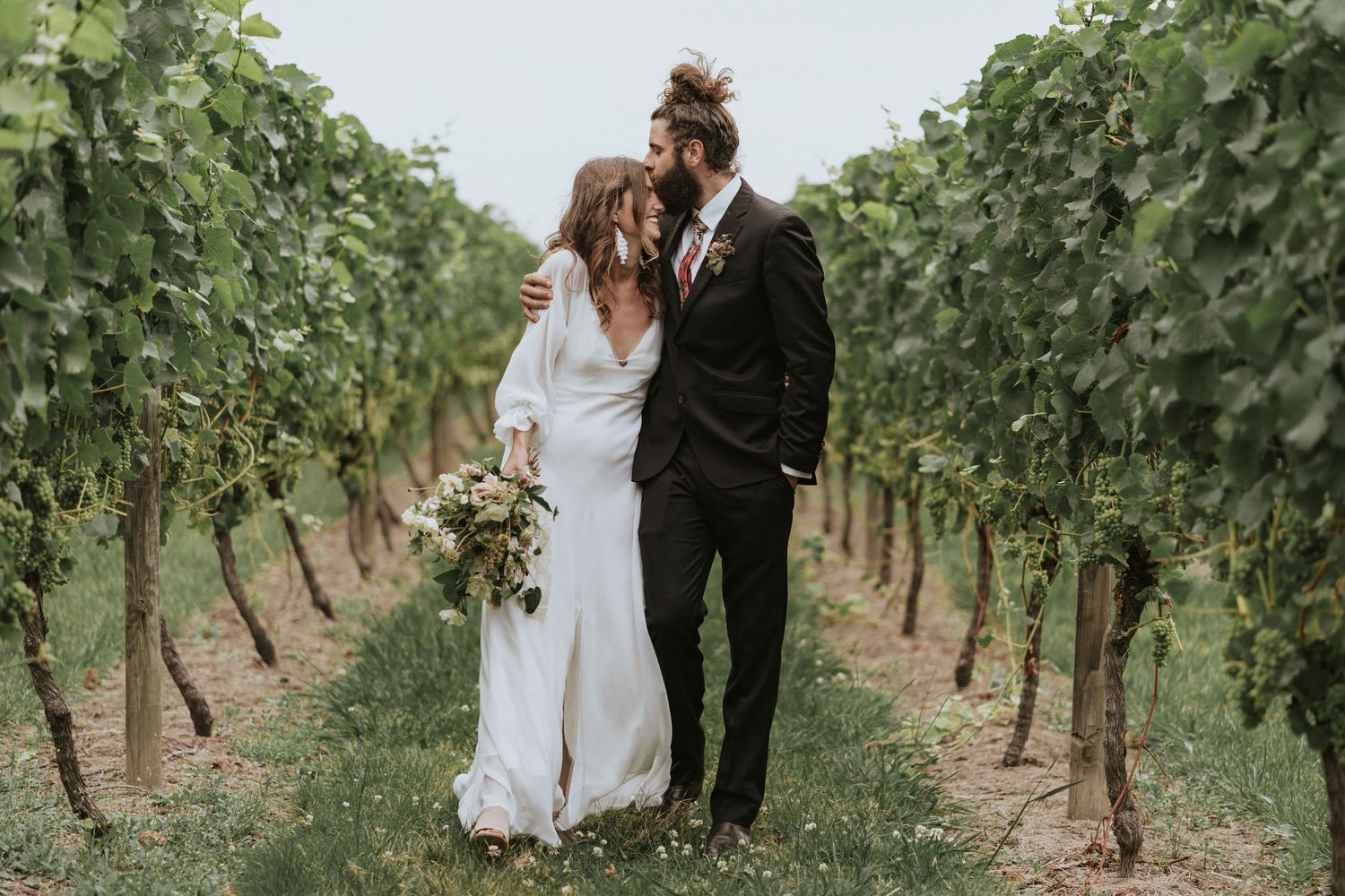 Couple walks intimately through lush green vineyard rows during romantic outdoor photoshoot session.