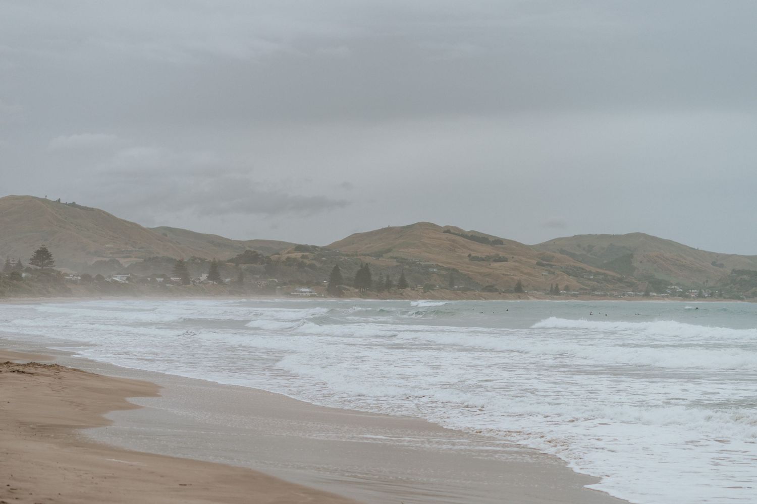 Moody coastal landscape with rolling hills and crashing waves on a foggy beach under overcast skies.