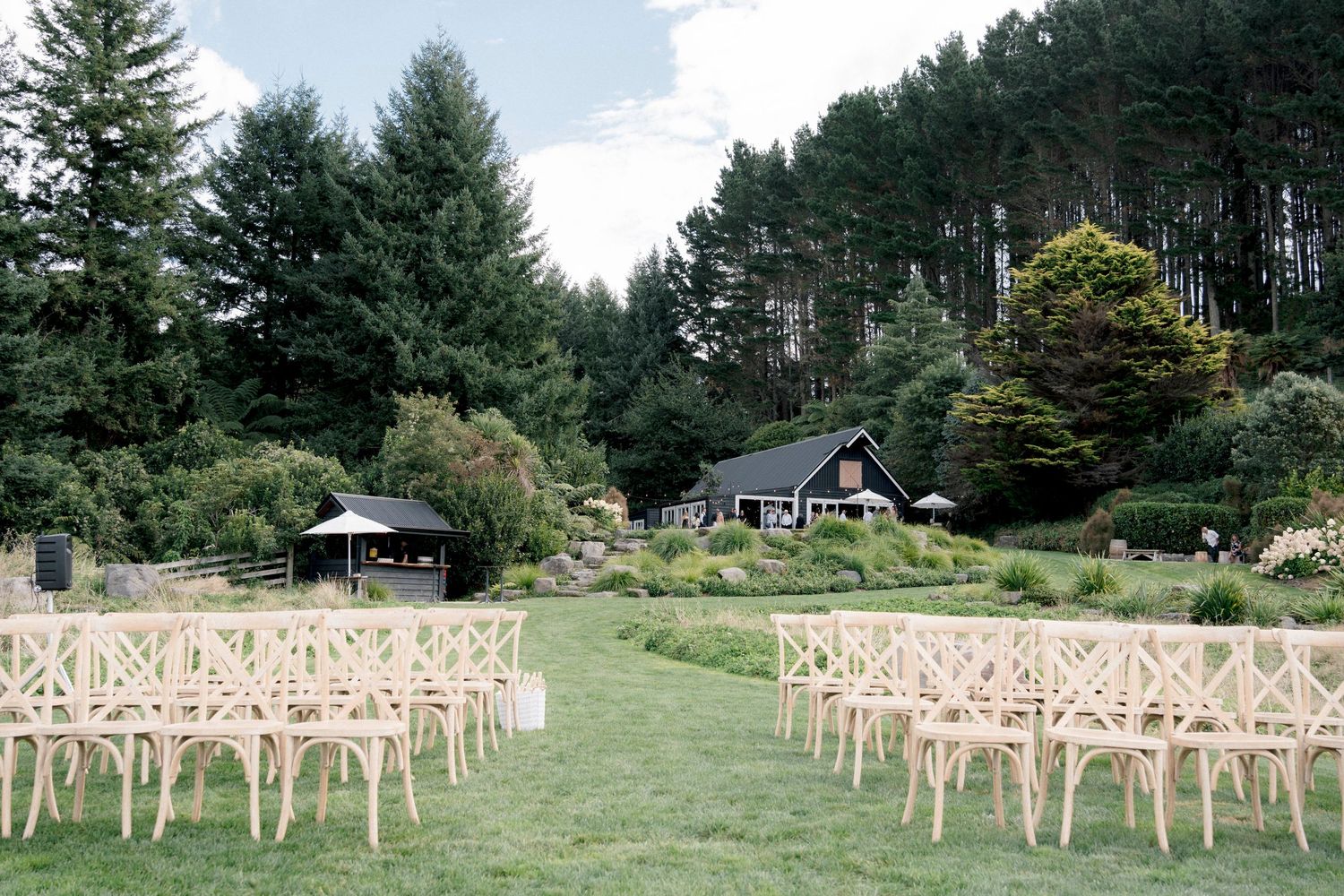 Rustic outdoor wedding ceremony setup with crossback chairs on a green lawn surrounded by evergreen trees and cabins.