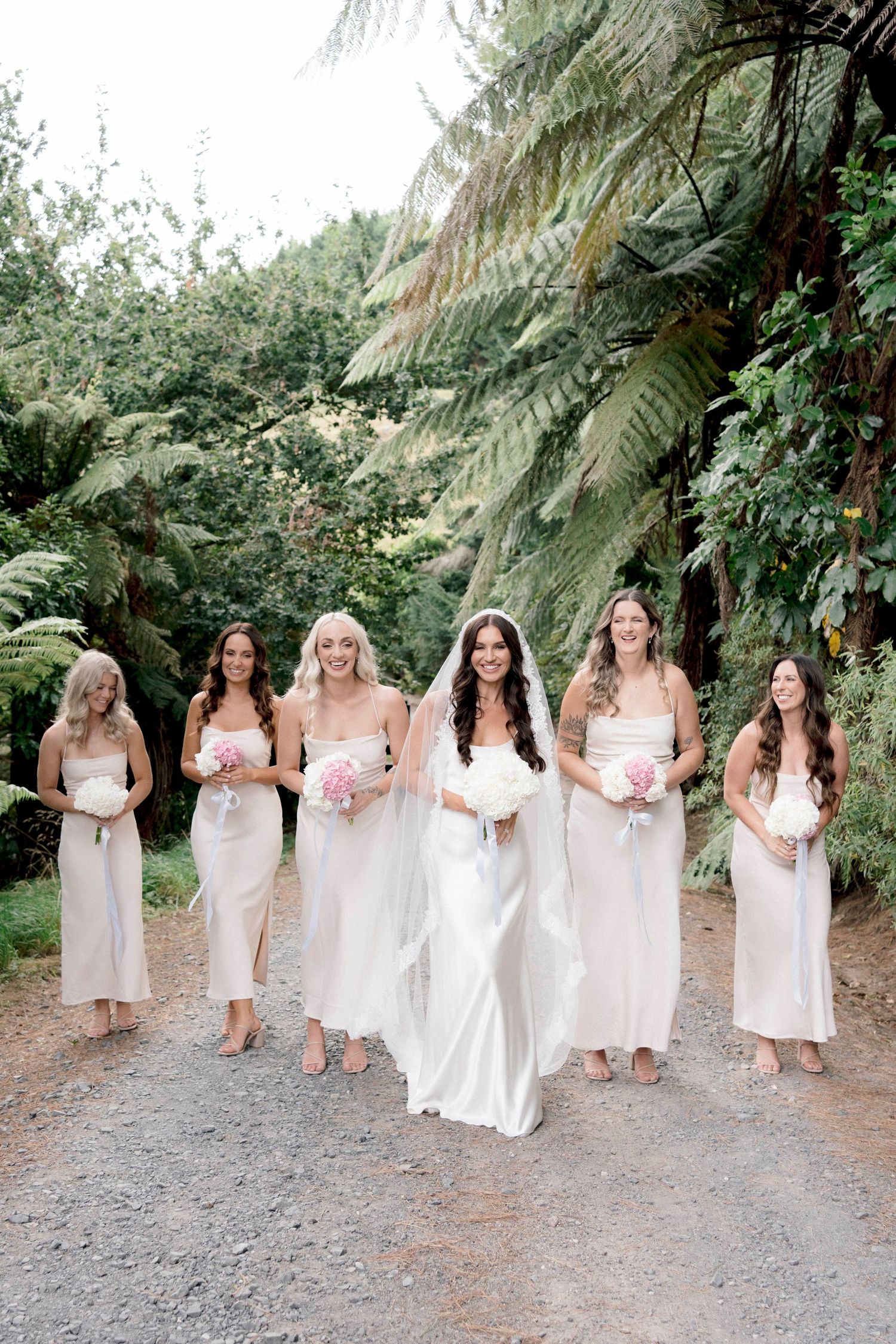 A bridal party in flowing white dresses poses on a path surrounded by lush ferns and greenery.