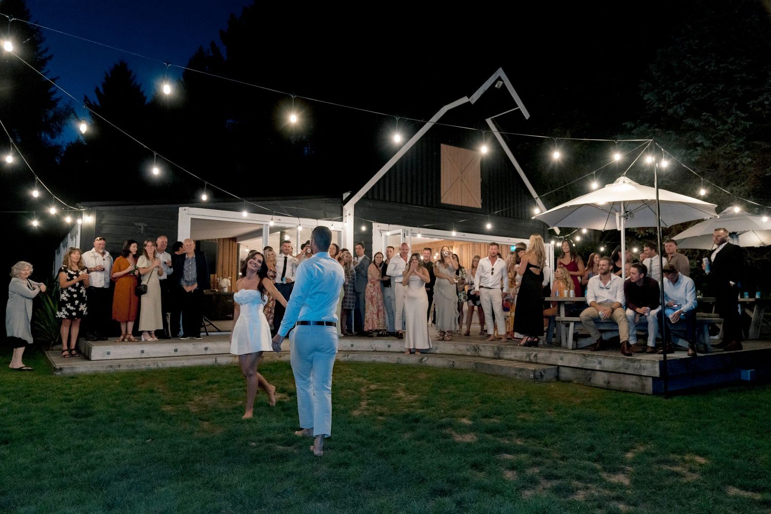 Evening wedding reception with string lights illuminating guests gathered outside a white building.