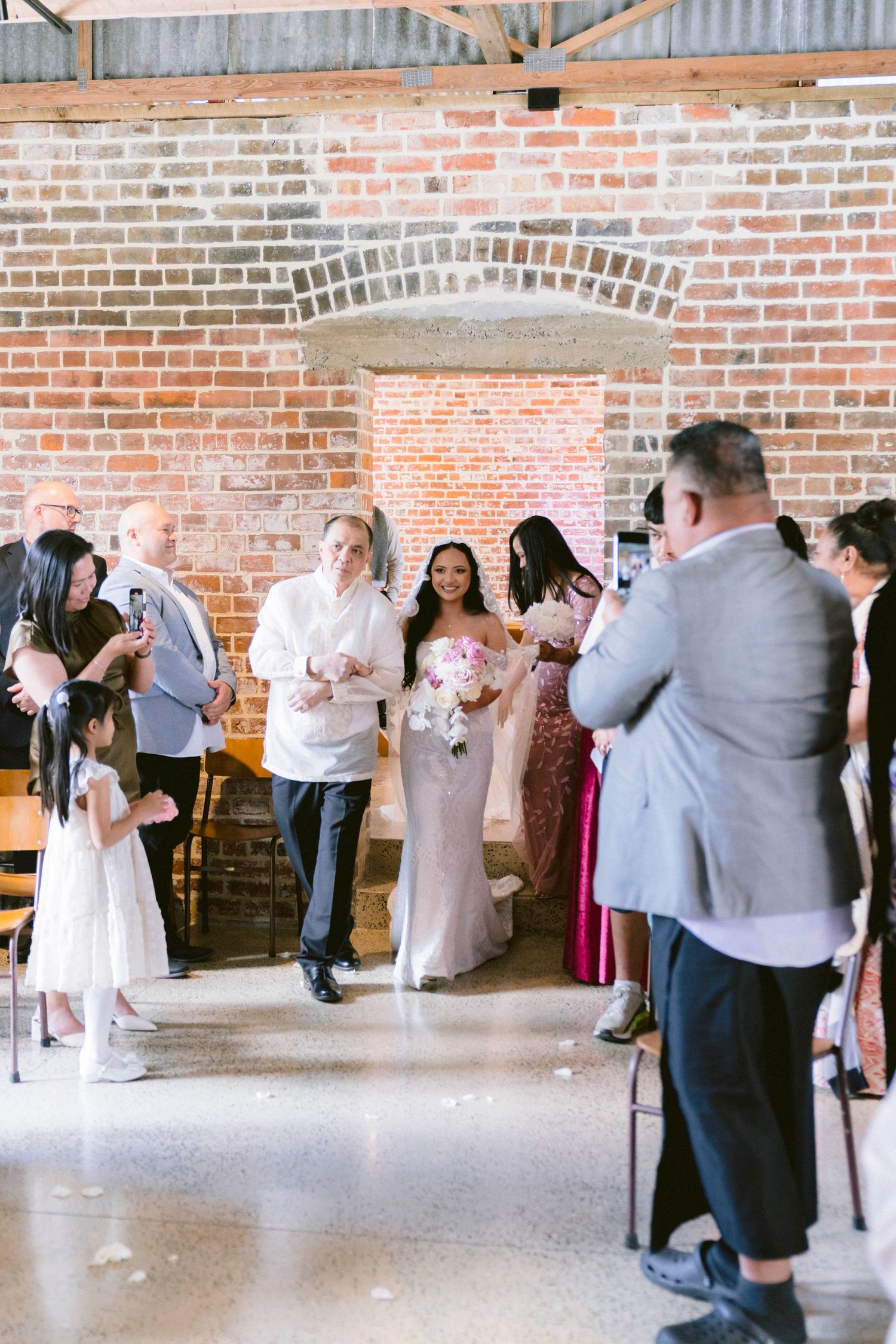 Wedding guests dance together in a rustic brick-walled venue with exposed beams and wooden chairs.