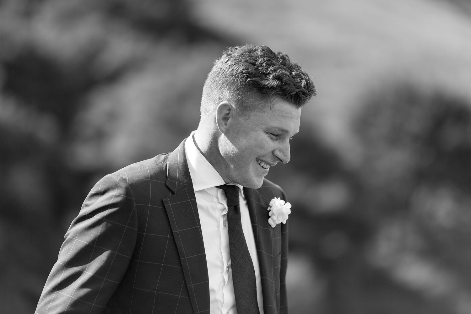 A person in a formal dark suit and white boutonnière smiles while looking to the side during what appears to be a wedding.