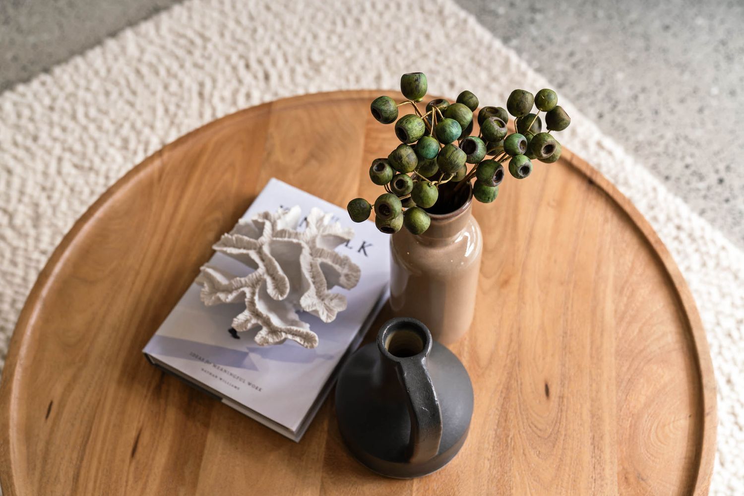 A wooden round table displays a hardcover book, a beige vase with green berries, and a small black ceramic vessel.