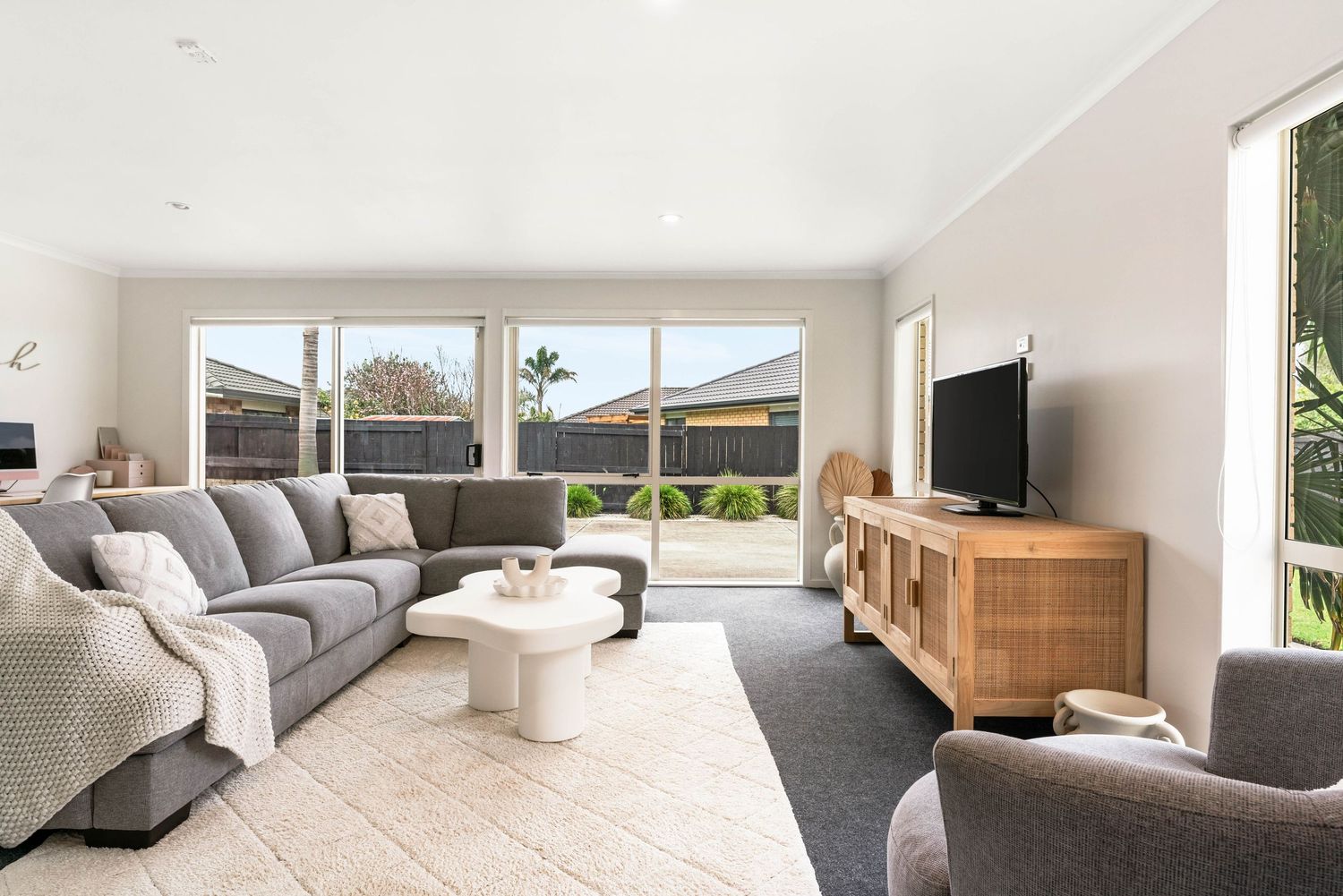 Modern living room interior with grey L-shaped sofa, wooden TV unit, and large windows overlooking the backyard.