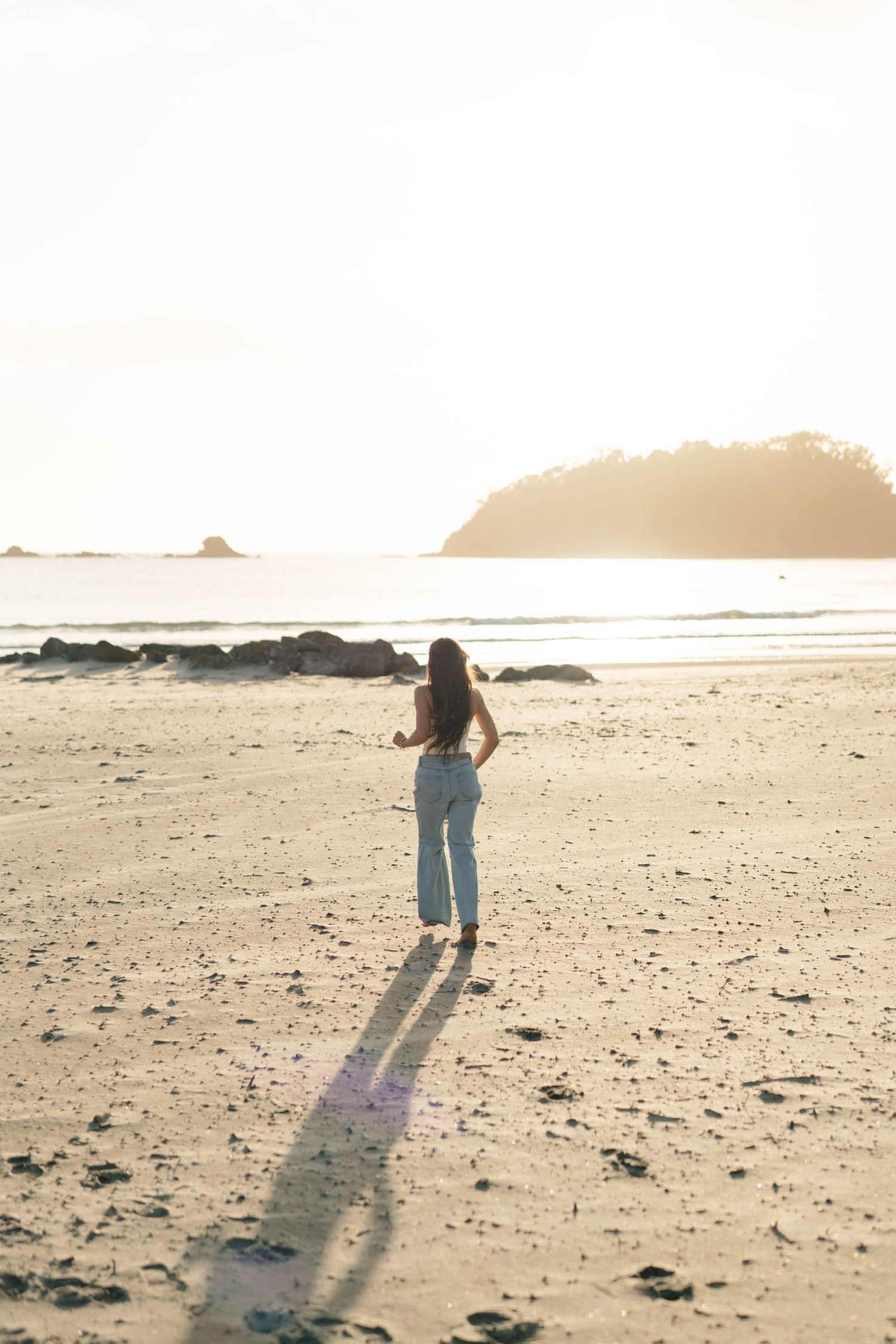 A person's silhouette walking along a misty beach at sunset casting a long shadow on the sand.
