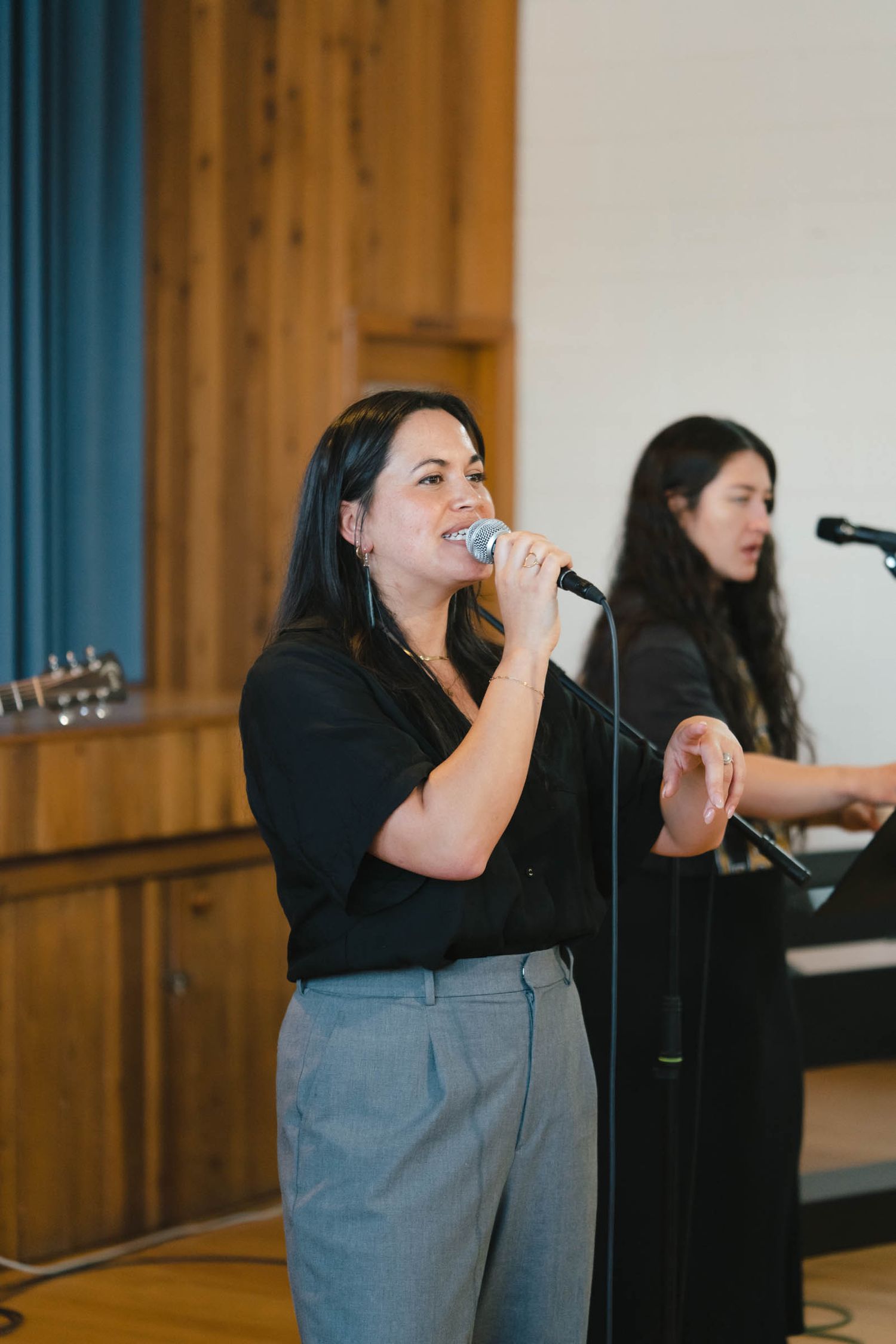 Two singers perform at a venue with microphones, one wearing black and the other in gray pants in a wooden-paneled room.