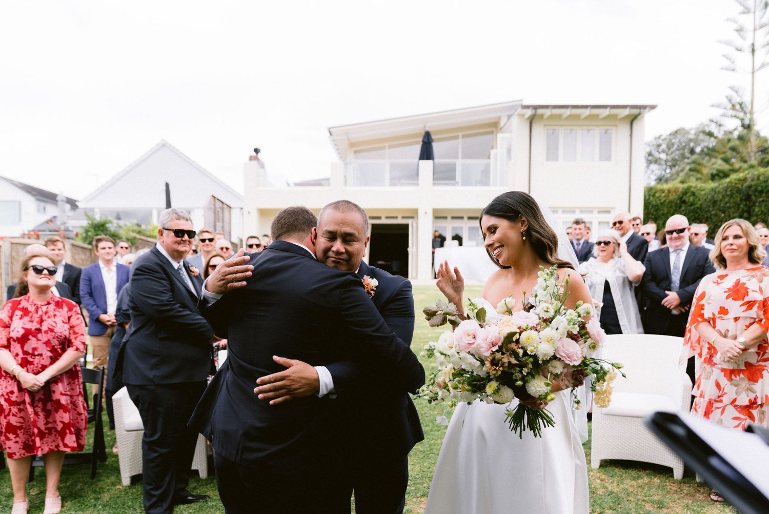 Wedding guests gather on a lawn for post-ceremony celebrations outside a white house.