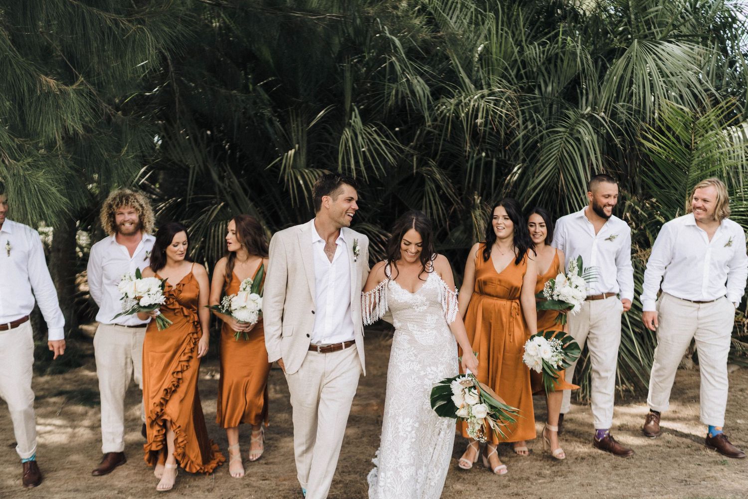 Wedding party walks outdoors wearing white and rust-colored attire among tropical palm trees.