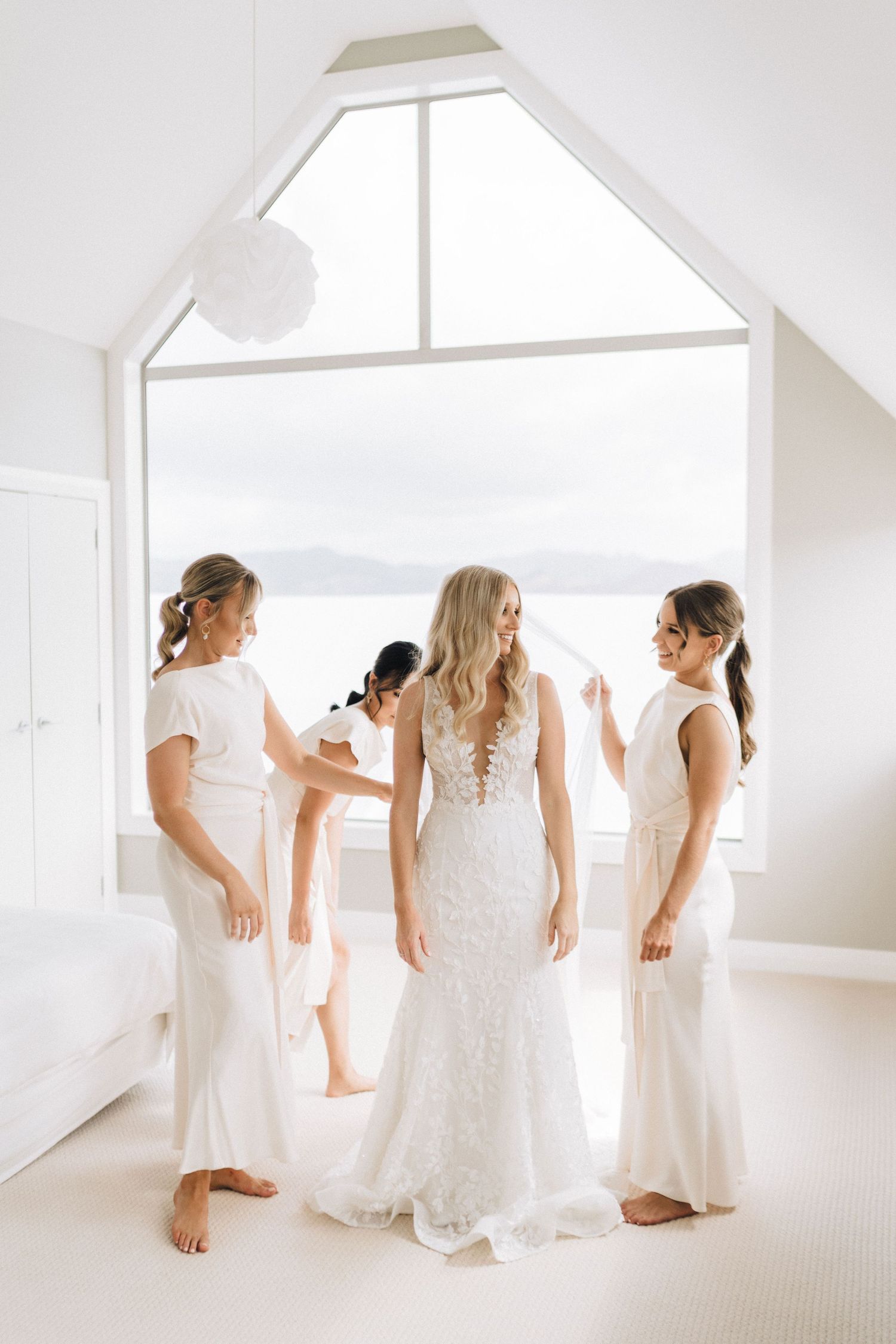 Three women in white dresses stand by a bright window overlooking mountains, preparing for a wedding celebration.