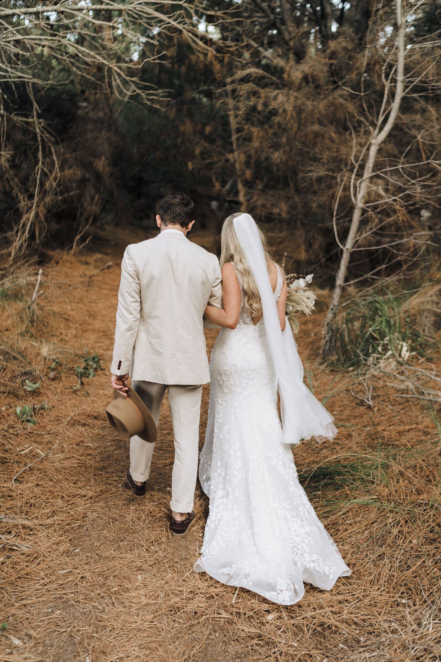 A couple walking along a dirt path in formal wedding attire with the bride wearing a flowing lace dress.