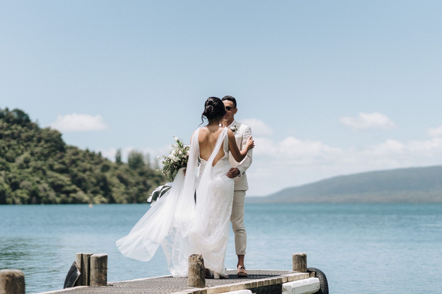 A wedding couple stands on a lakeside pier with mountains in the background as the bride's dress flows in the wind.