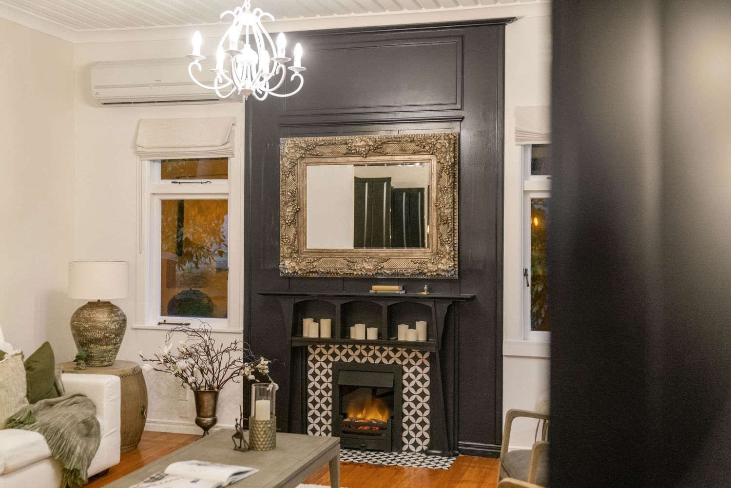 Elegant living room with black accent wall, ornate gold mirror, and crystal chandelier above a decorative fireplace.