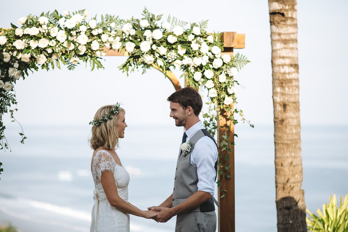 Romantic wedding ceremony setup with white floral decorations and tropical foliage against an ocean backdrop.