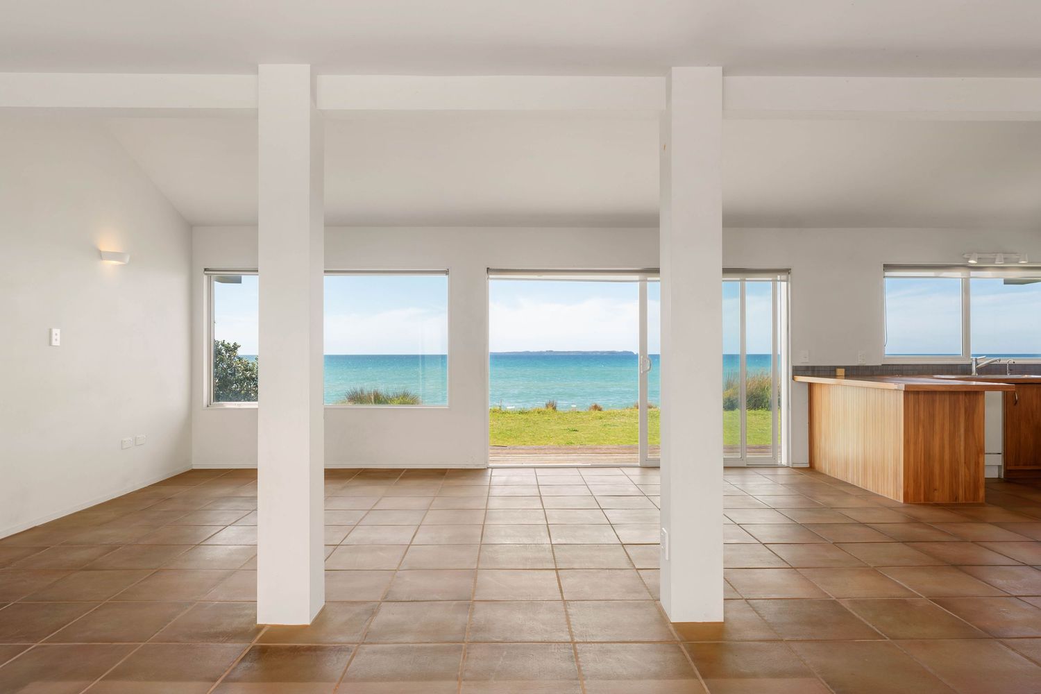 Minimalist interior space with white columns and tiled flooring offering expansive views of the ocean and lawn.