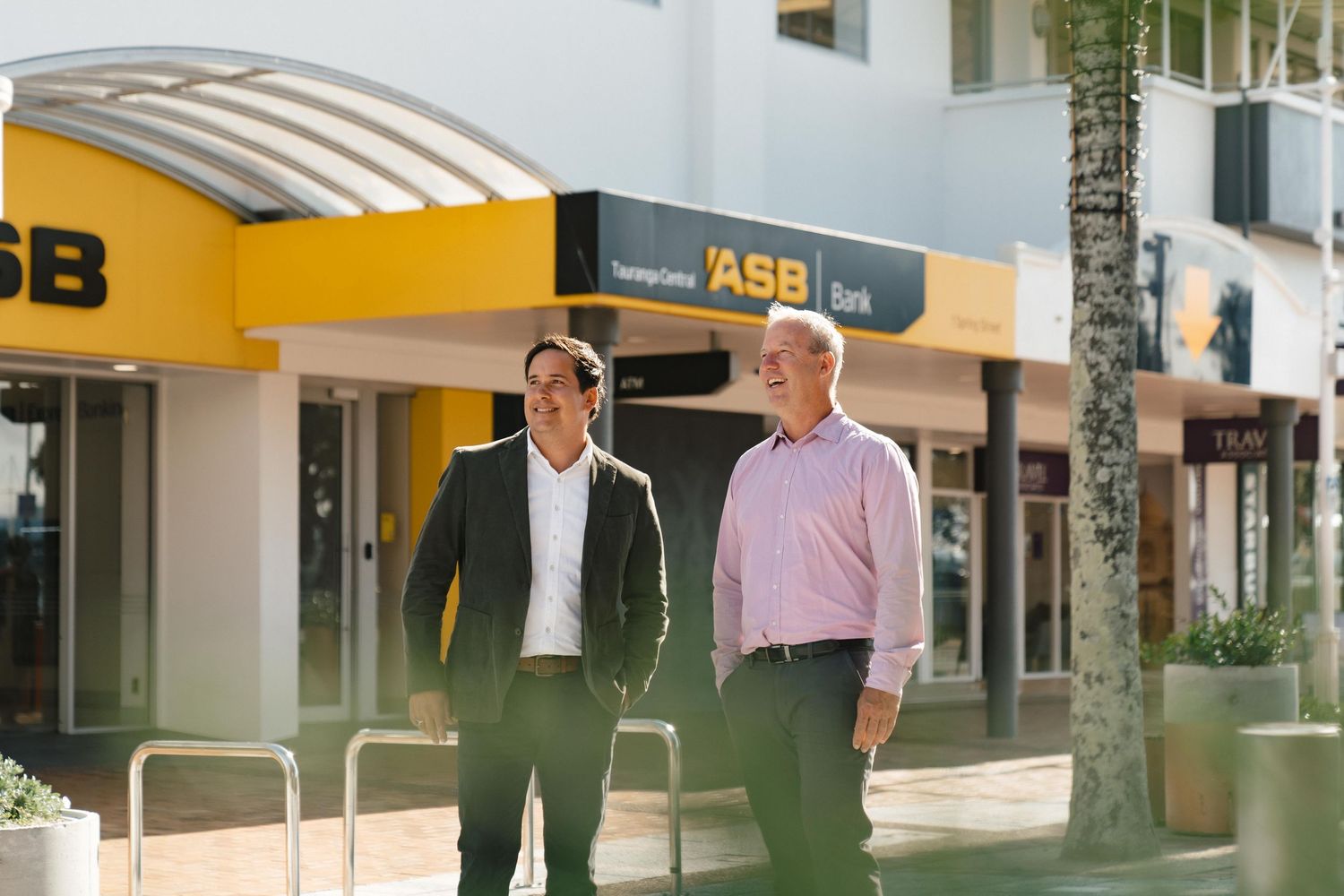 Two professionals walking outside ASB Bank branch with yellow awning and palm trees.