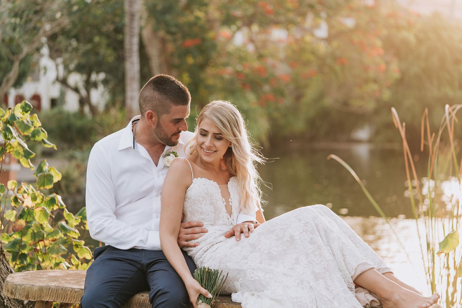 A romantic couple sits by a pond at sunset with the bride wearing a white lace dress.