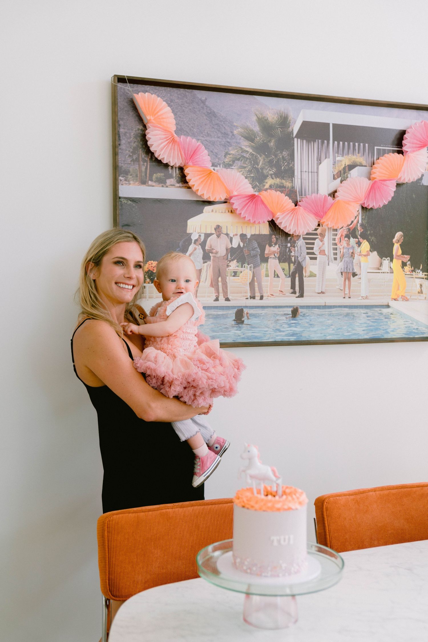 Birthday celebration with pink decorations including a cake on stand and coral paper garland hanging on wall above.